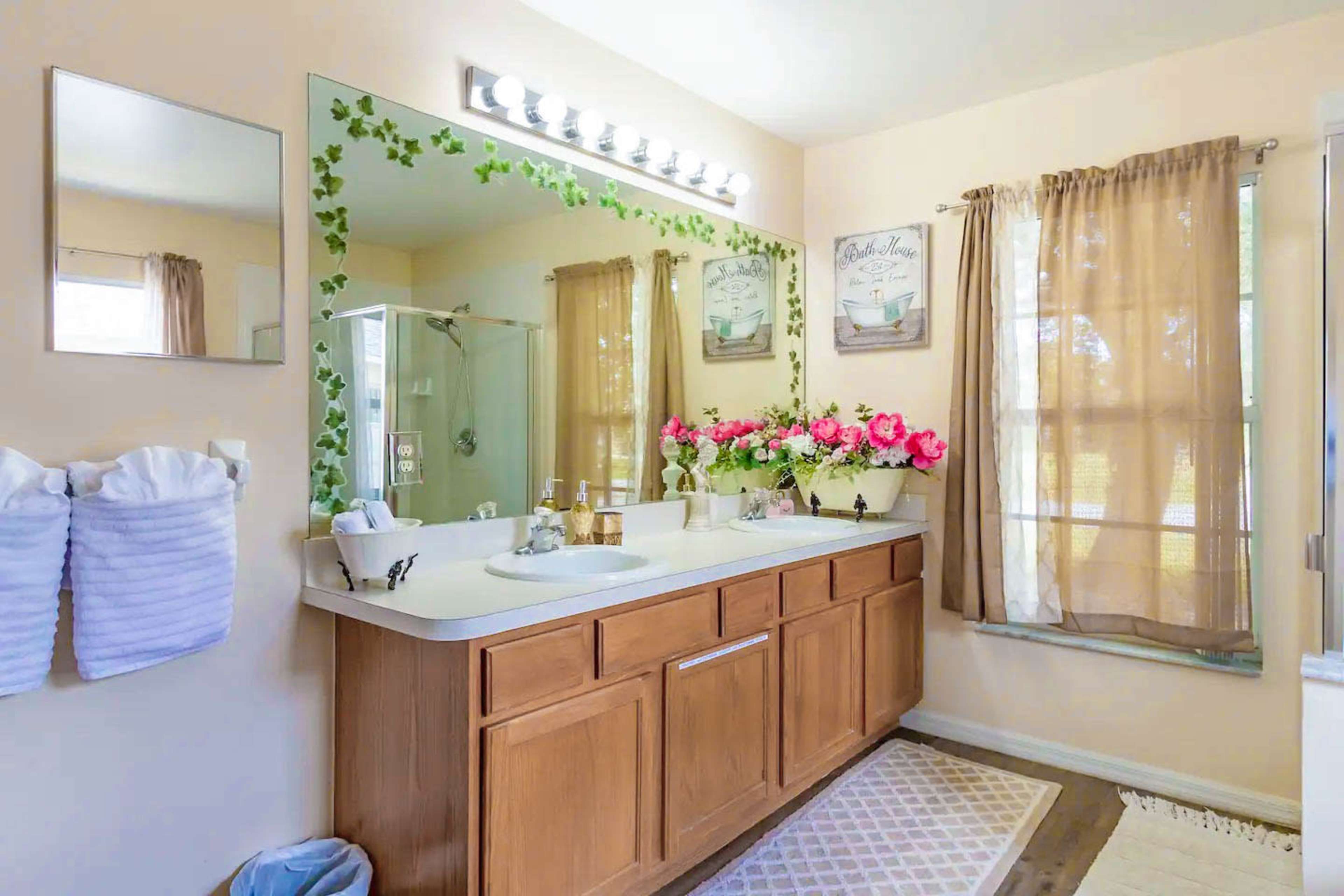 A bathroom with a wooden double vanity, a large mirror above it, and curtains on the windows, complemented by floral decorations on the countertop.