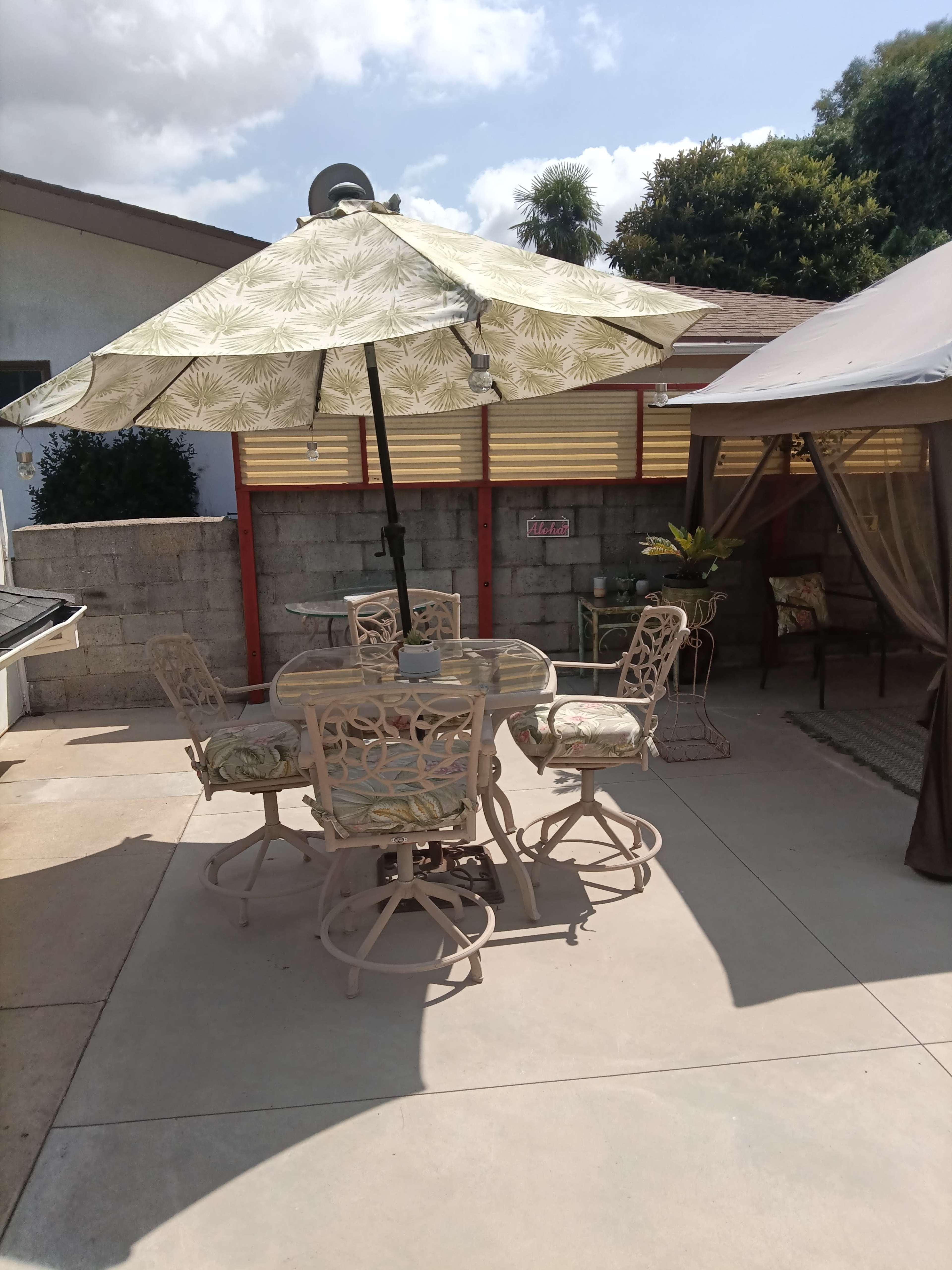A patio area features a round table with four tall chairs under a large umbrella, alongside a shaded seating area with a canopy.