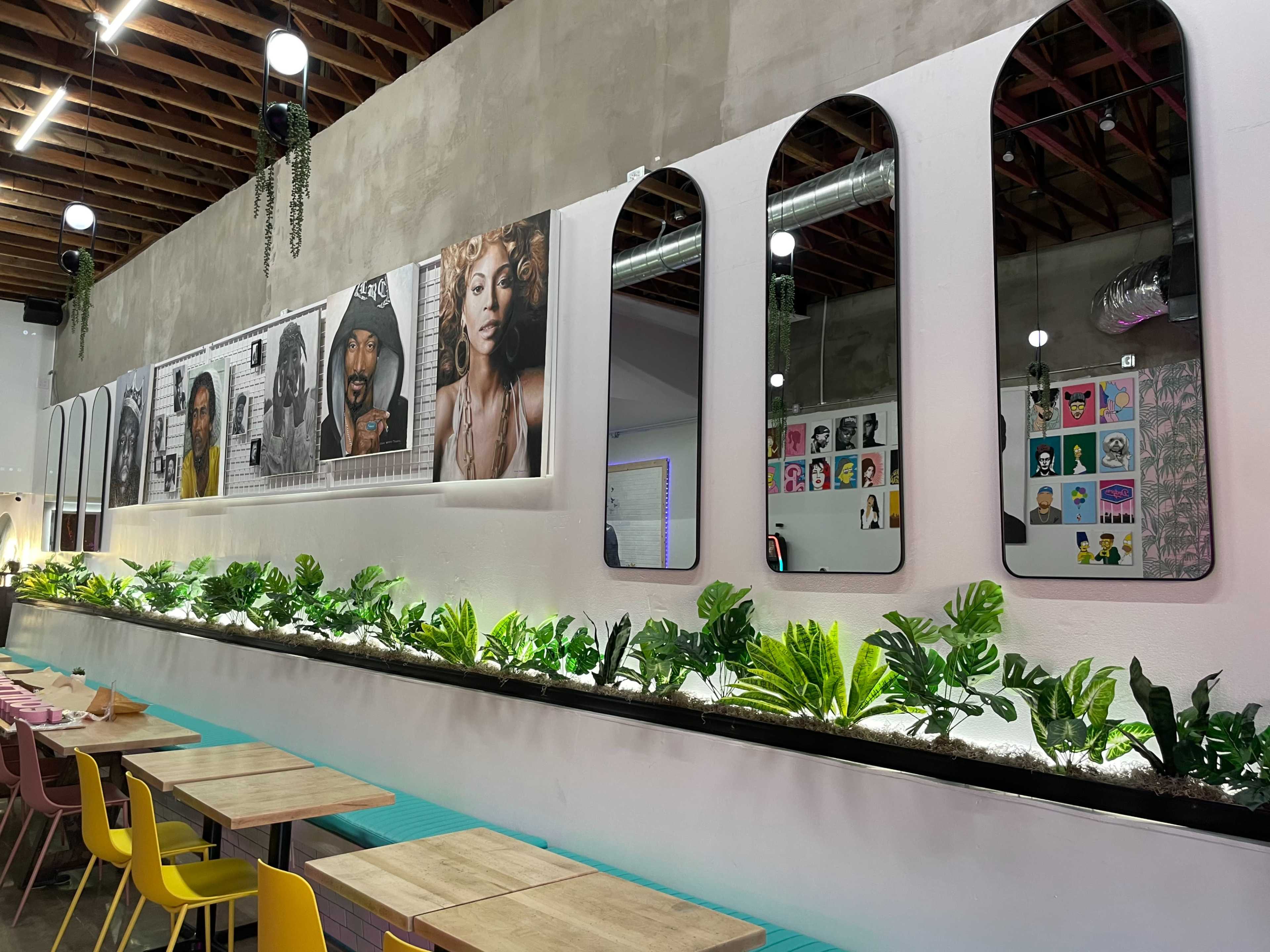 The image shows a cafe interior with a wall displaying framed artworks and mirrors, and a row of green plants along the bottom.