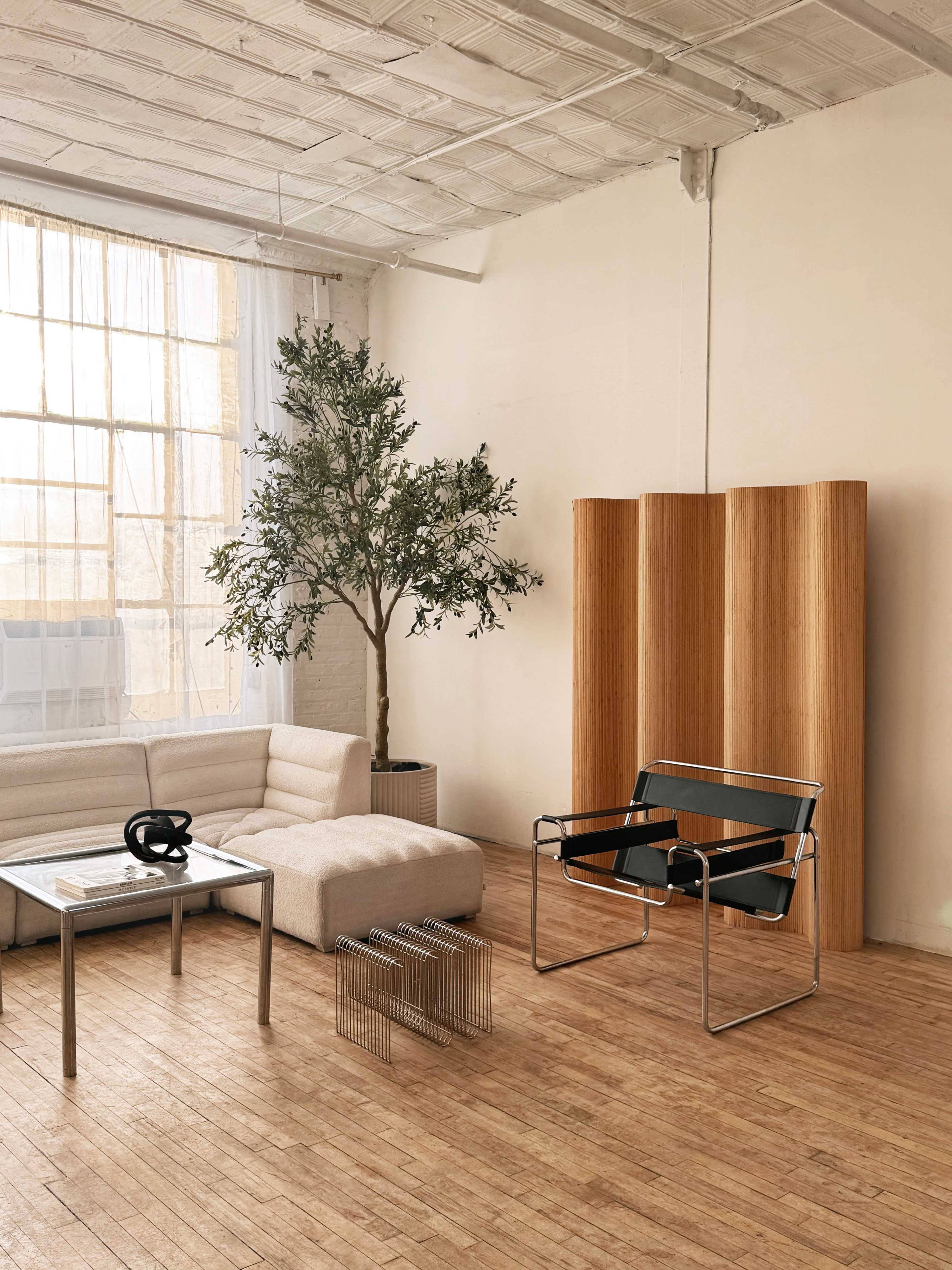 The image shows a minimalist living space with a light-colored sofa, a glass coffee table, a metal chair, and a potted tree, all set against a backdrop of large windows and wooden flooring.