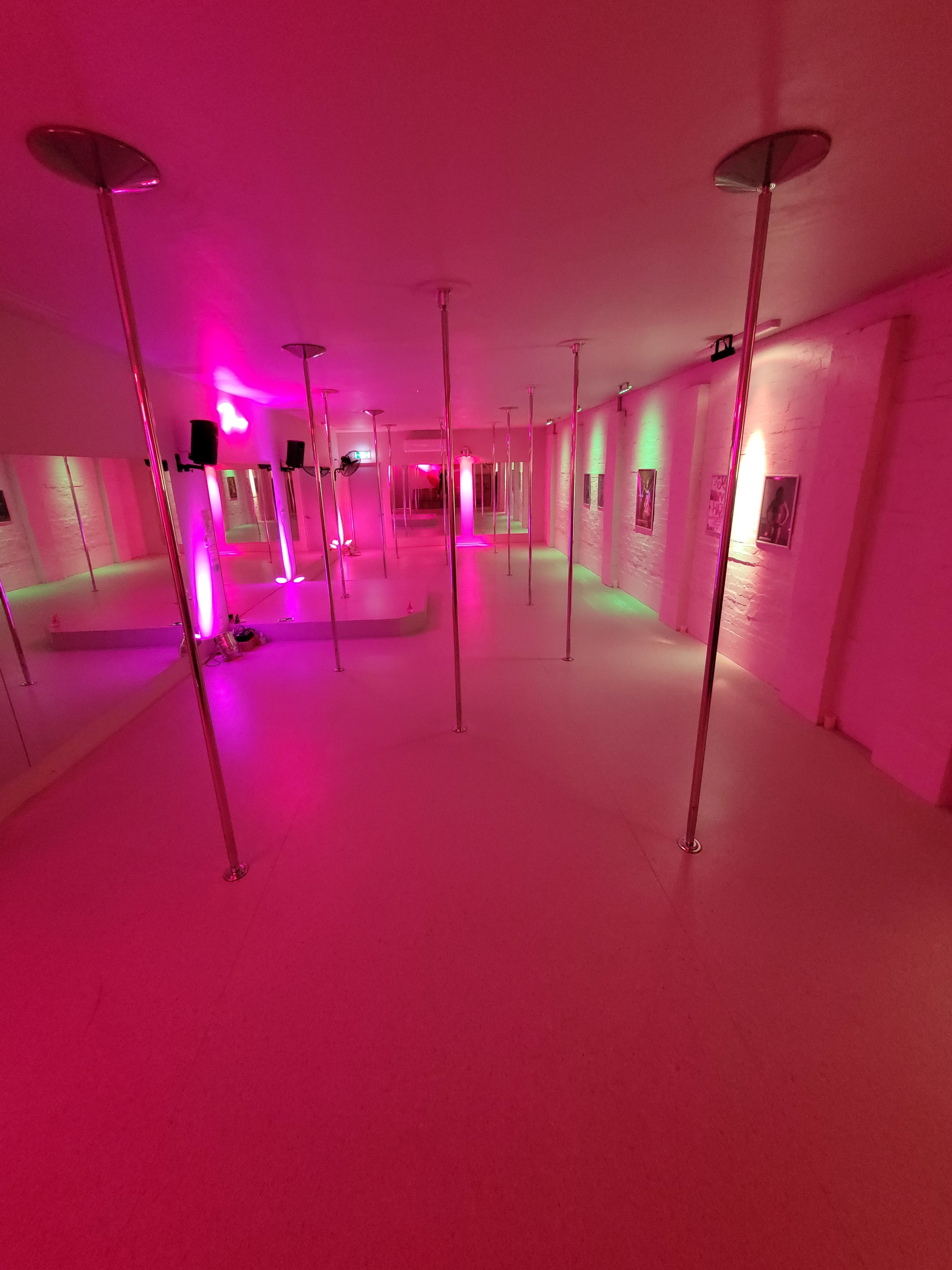 The image shows a spacious dance studio featuring multiple mirrored walls and several polished dance poles illuminated by pink and green lights.