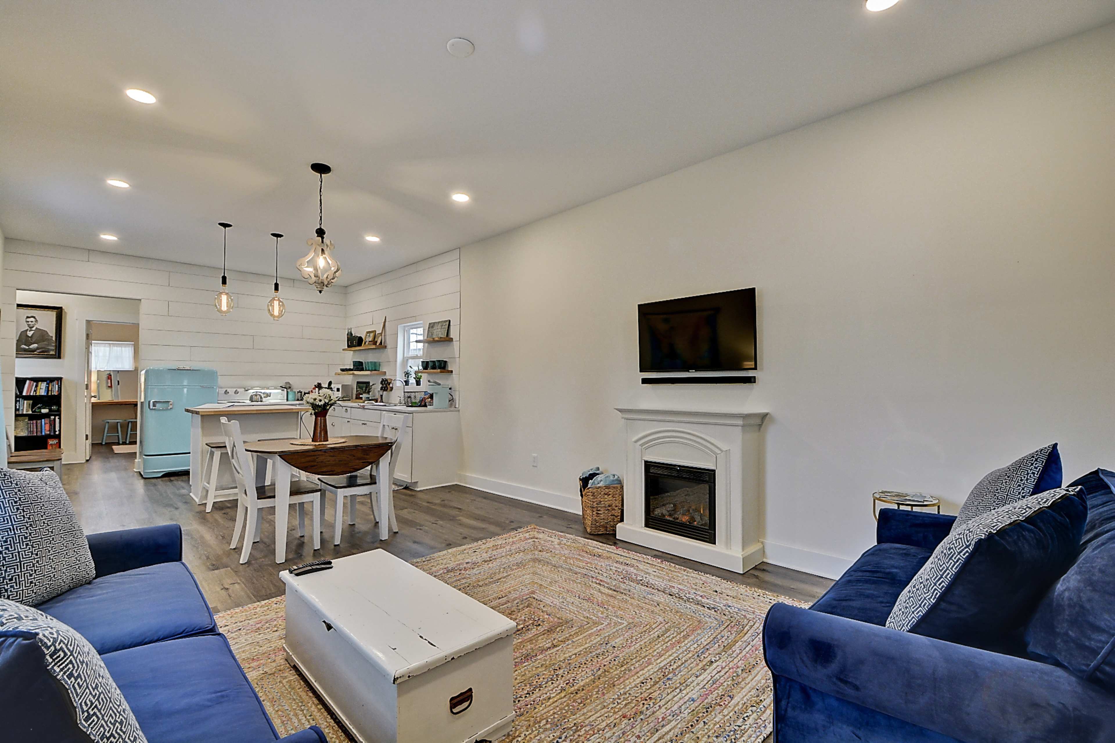 The room features a cozy living area with blue sofas, a white fireplace, and a dining space with a round table amid light-colored walls and wooden flooring.