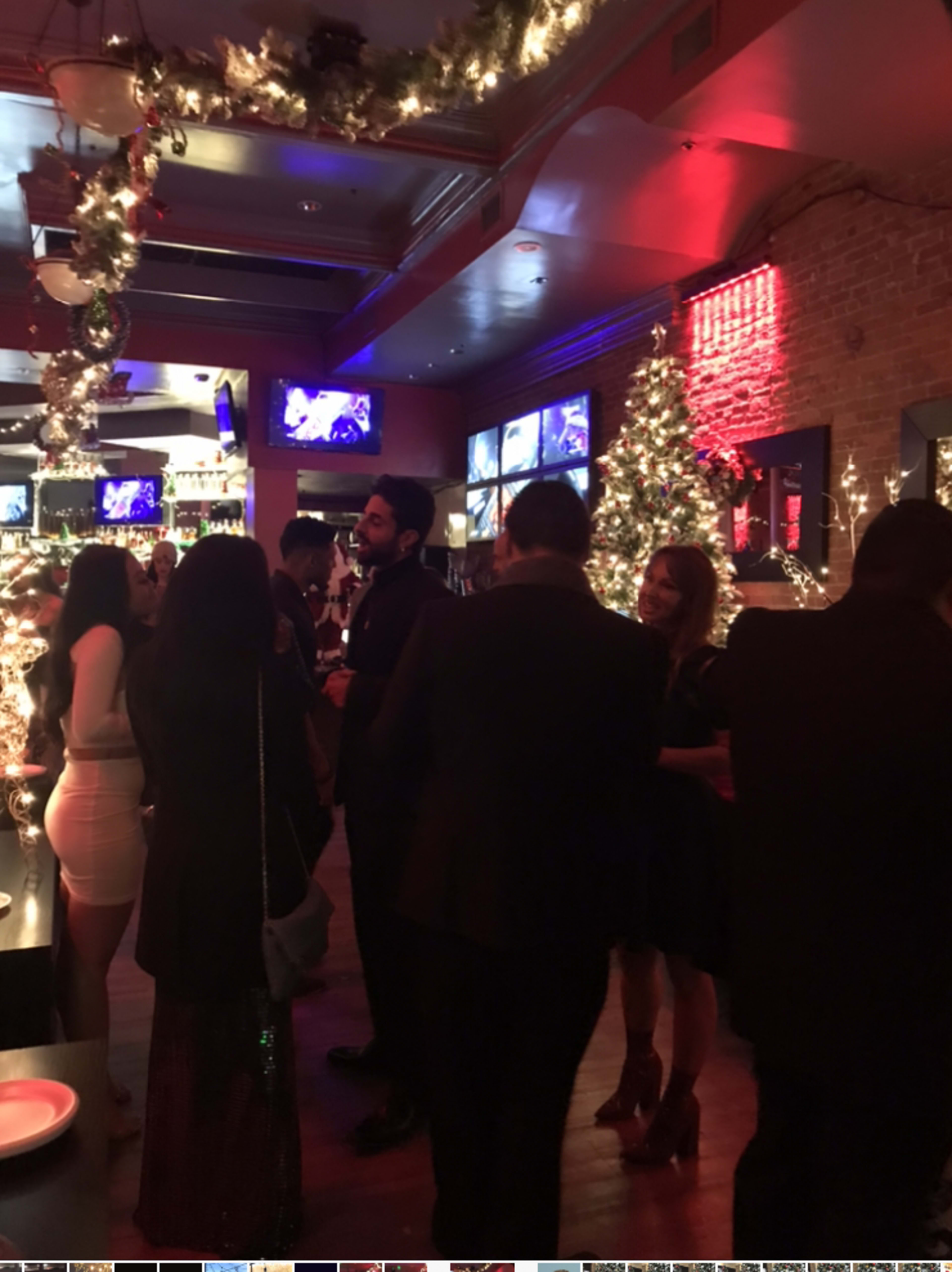 A festive bar scene features patrons mingling under holiday decorations, illuminated by colorful lights and multiple screens displaying entertainment.