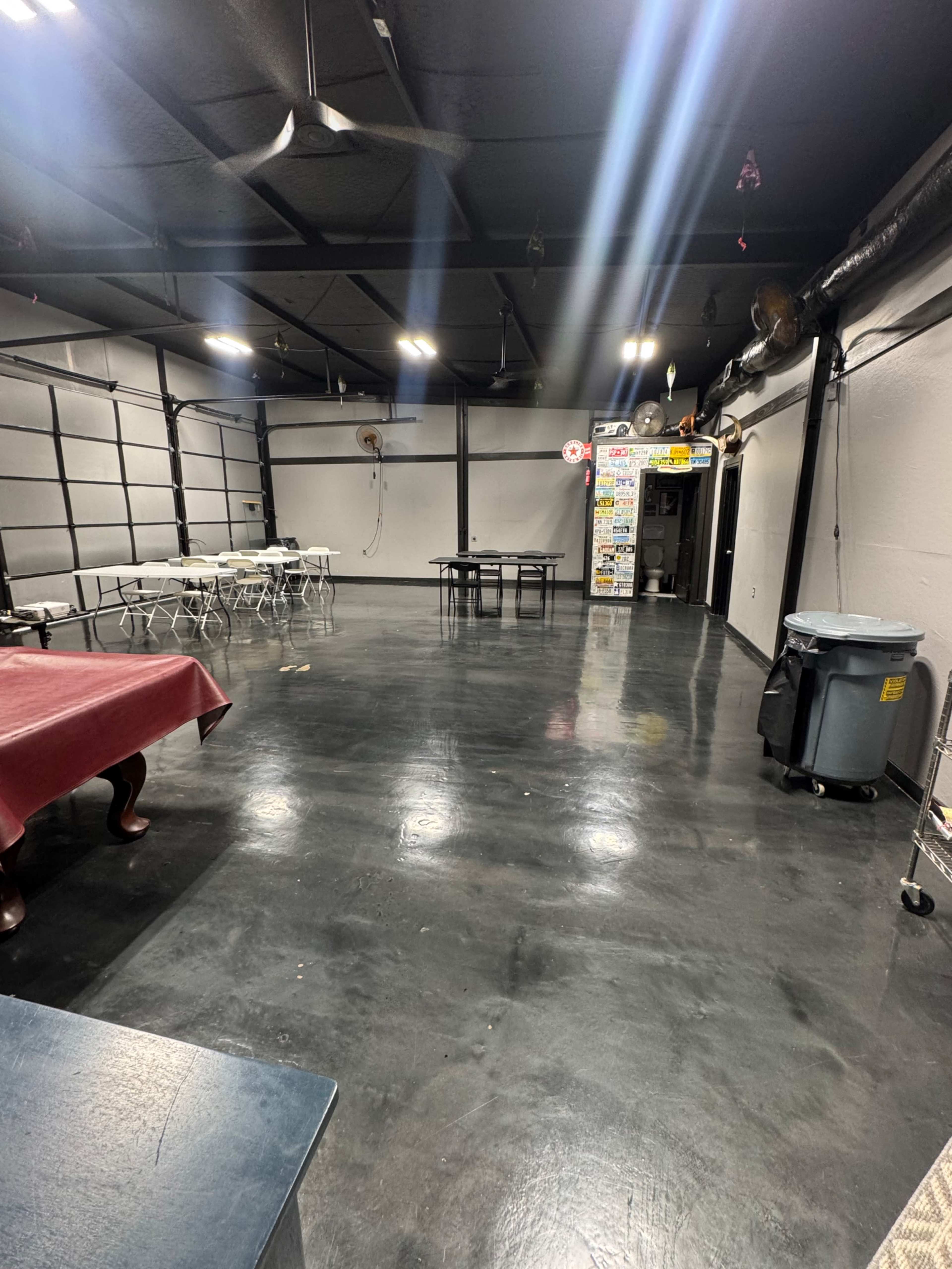The image shows a large, empty room with a polished concrete floor, a few tables and chairs on one side, and walls with a neutral color.