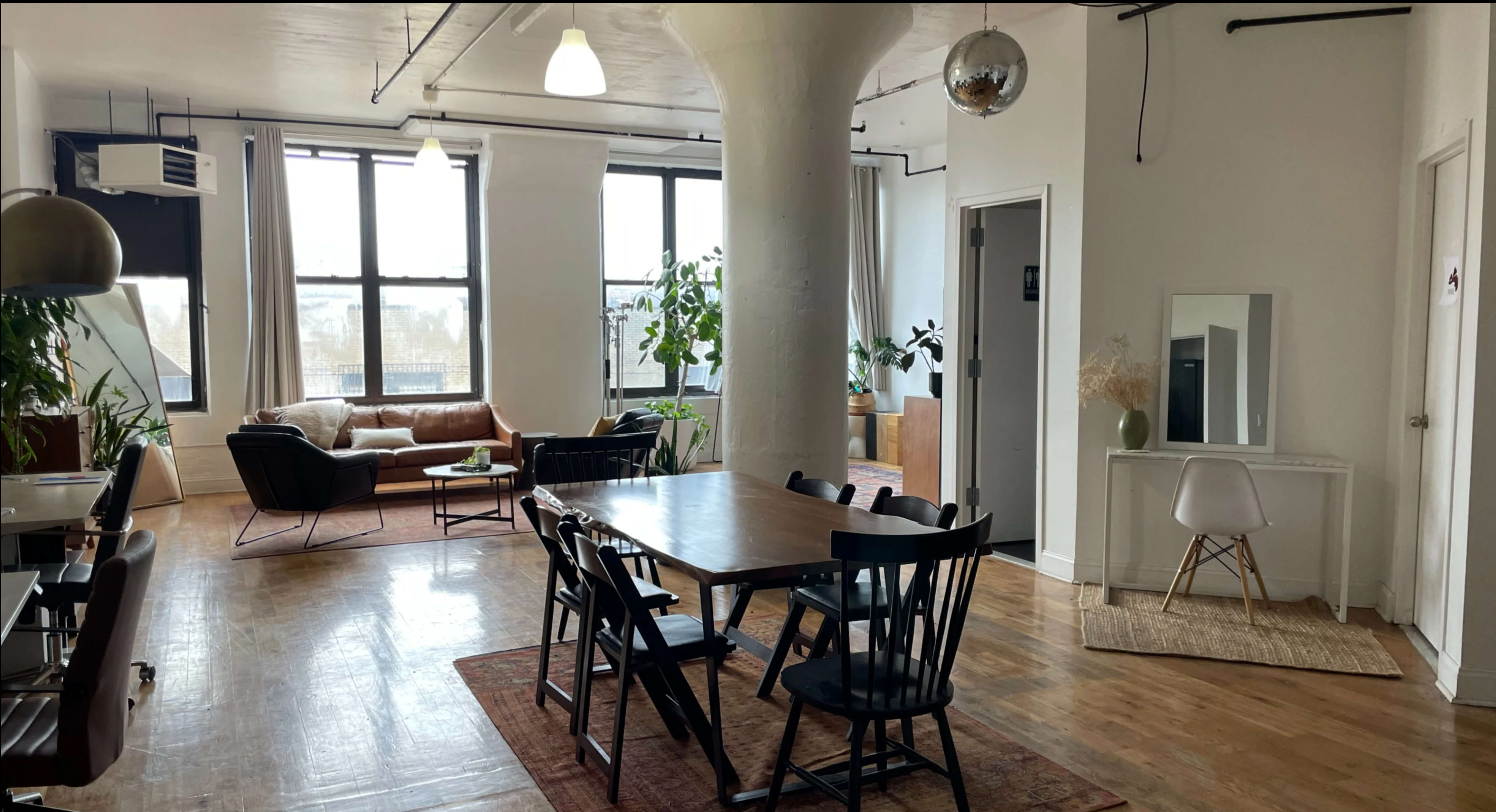 The image shows a spacious, well-lit open-concept living area featuring a dining table with chairs, a leather couch, large windows, and indoor plants.