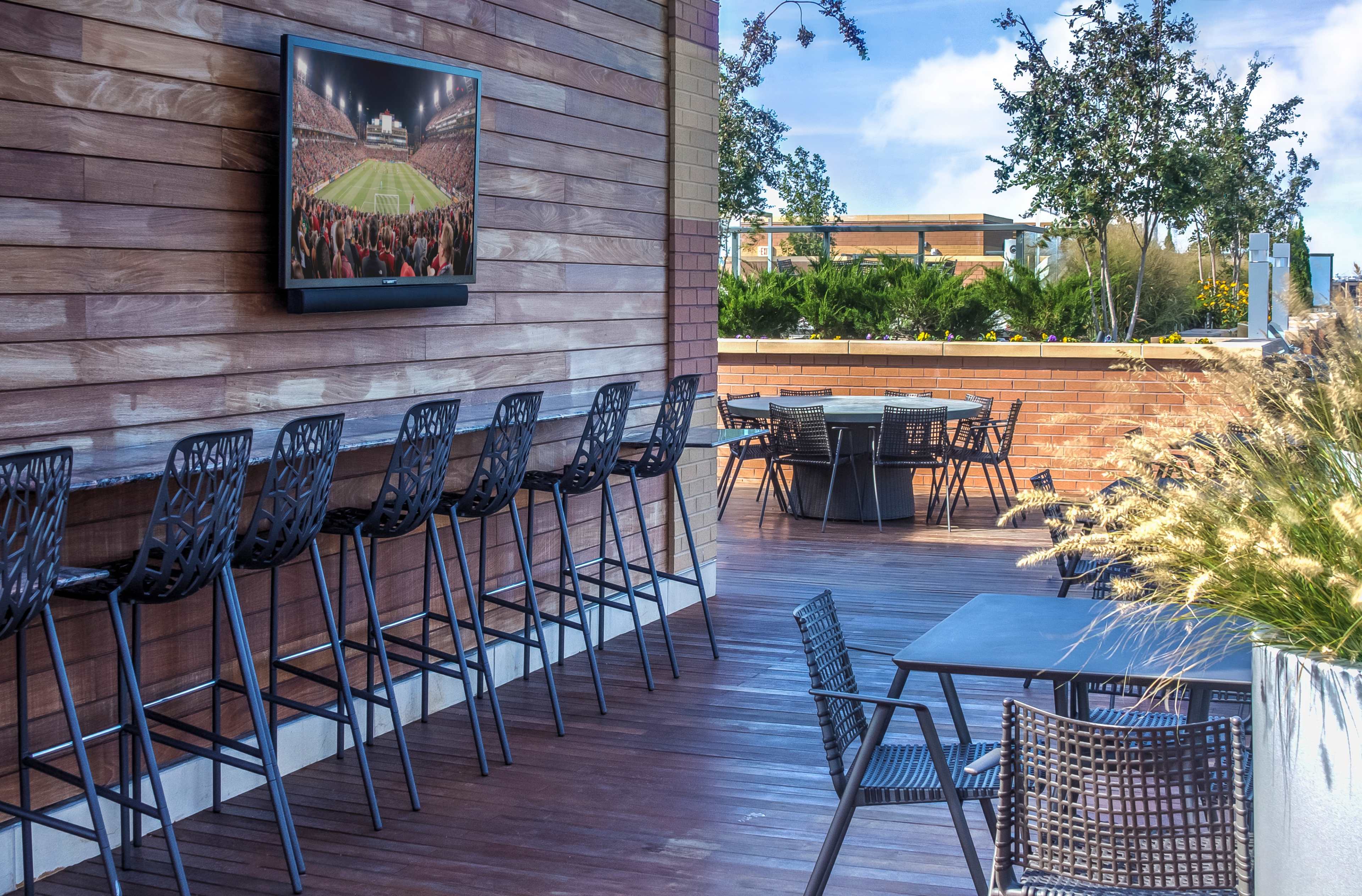 A modern outdoor seating area with wooden walls, high bar stools, a flat-screen television mounted on the wall, and tables arranged on a wooden deck surrounded by greenery.