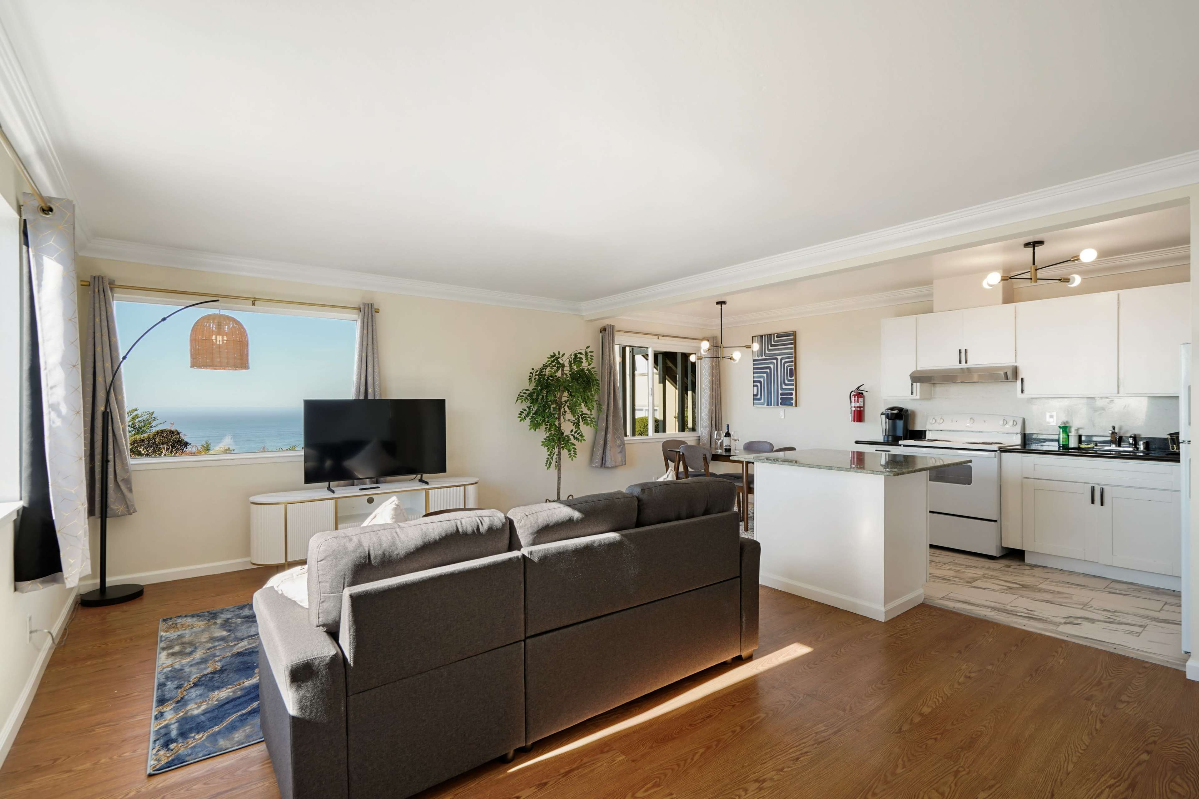 The image shows a modern living room and kitchen area with a large window offering a view of the ocean.