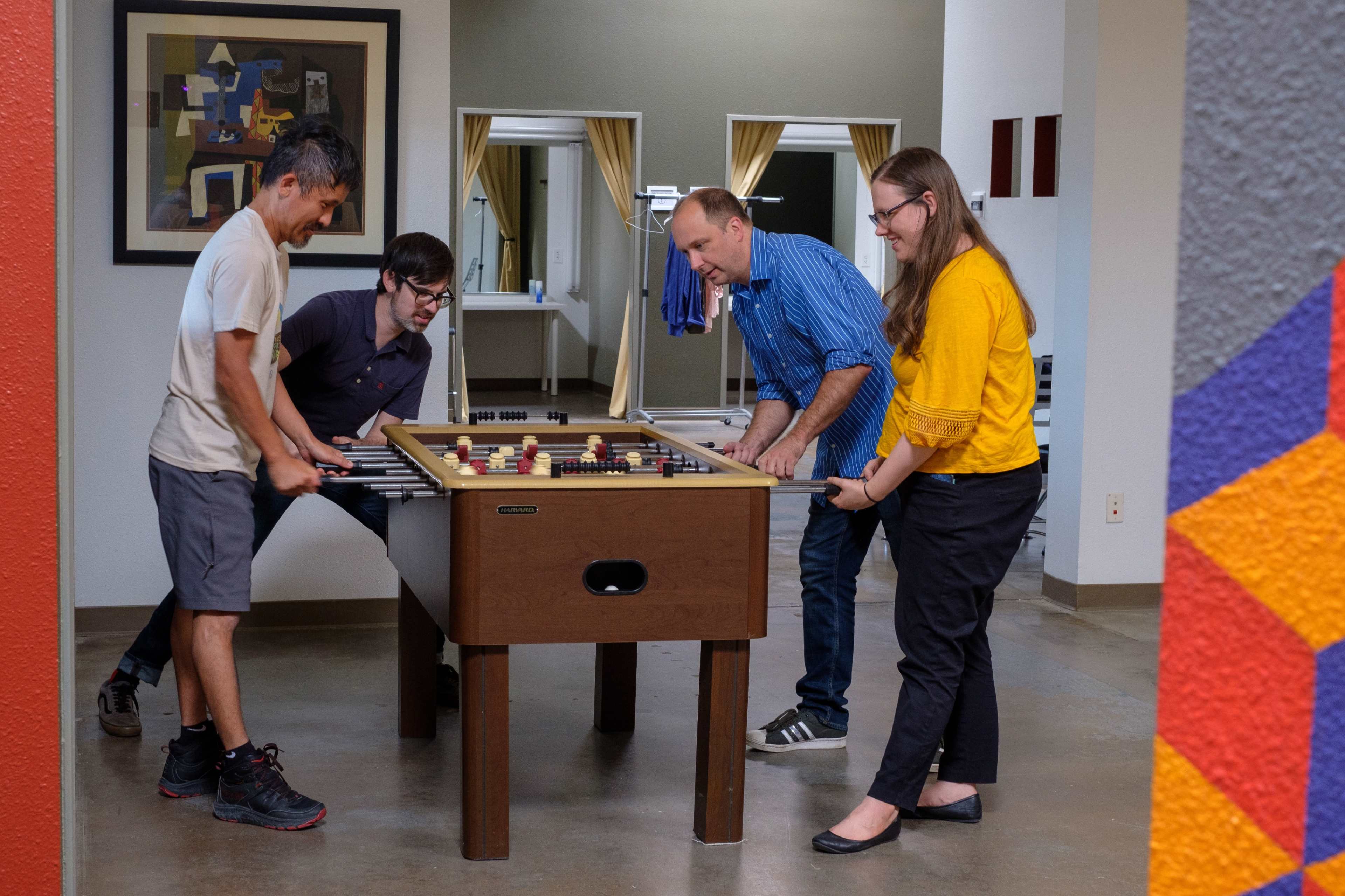 Four people are playing foosball in a brightly lit room with artwork on the walls.