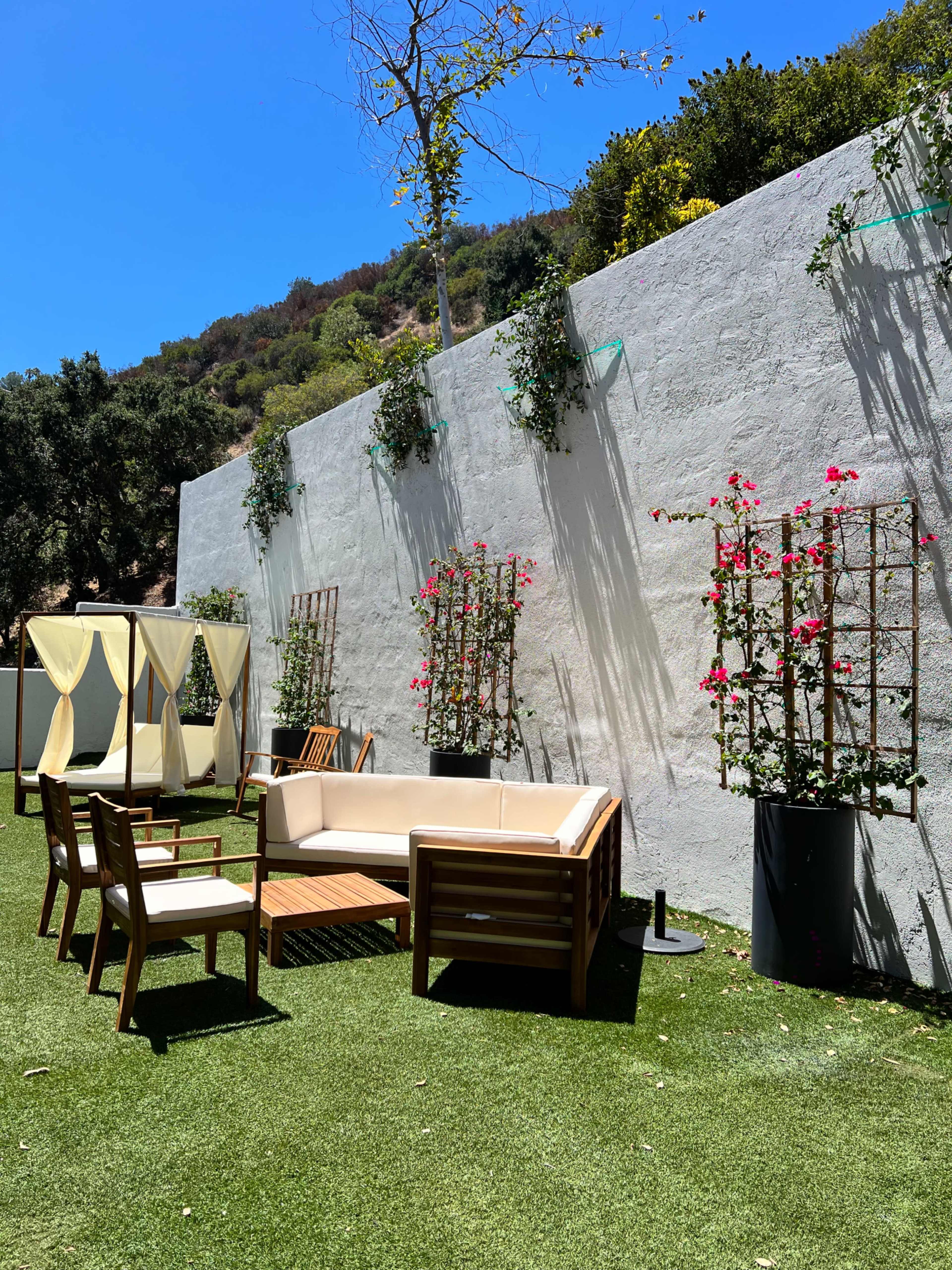 The scene features a outdoor lounge area with a beige sofa, wooden chairs, and decorative plants against a white wall, surrounded by lush greenery under a clear blue sky.