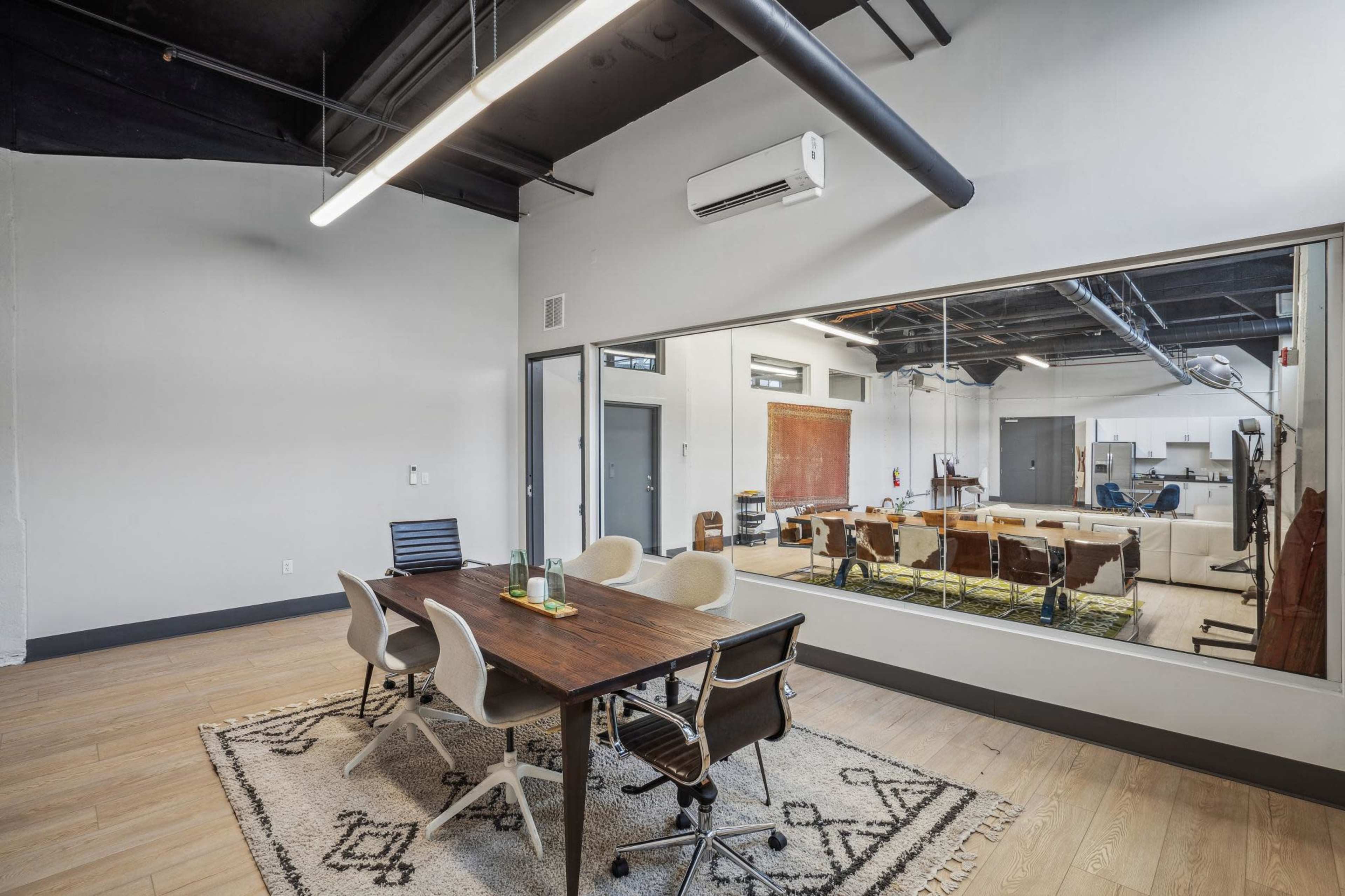 A conference room featuring a wooden table surrounded by modern chairs, with a large window overlooking an open office space equipped with various workstations.