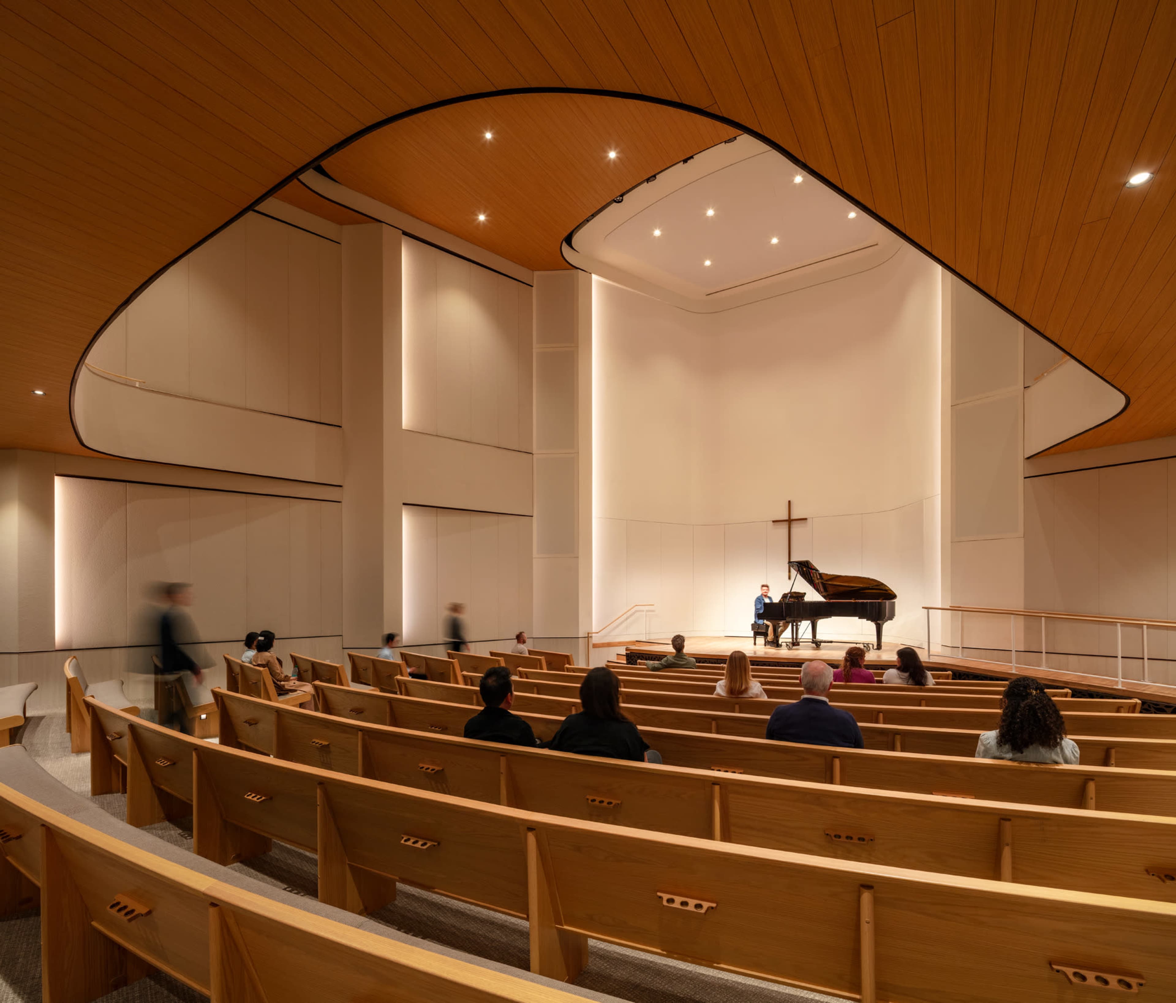 A pianist performs on stage in a modern chapel filled with wooden pews and a large cross in the background.