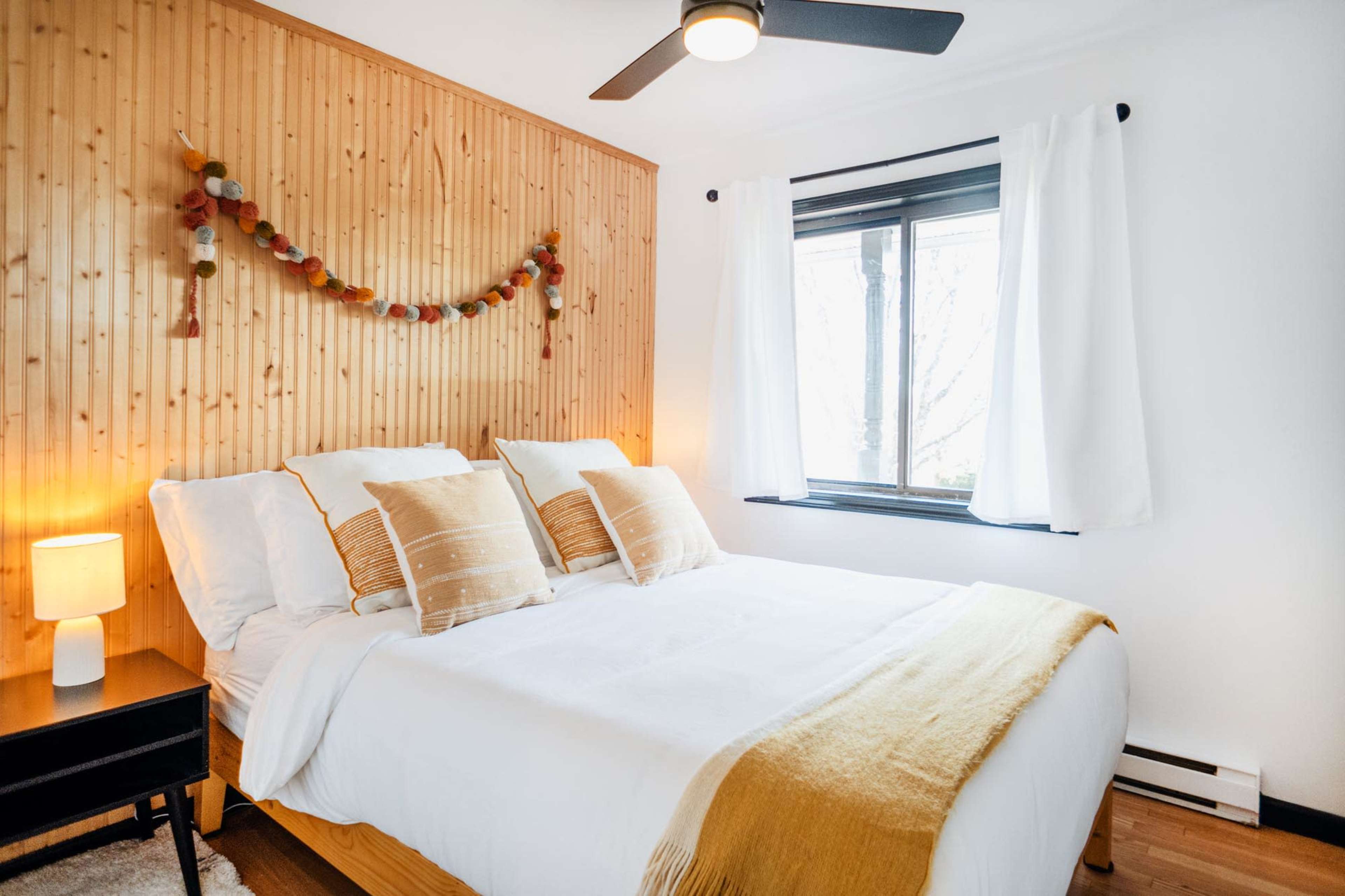 A neatly arranged bedroom with a bed featuring white linens, decorative pillows, and a wooden accent wall adorned with a garland.