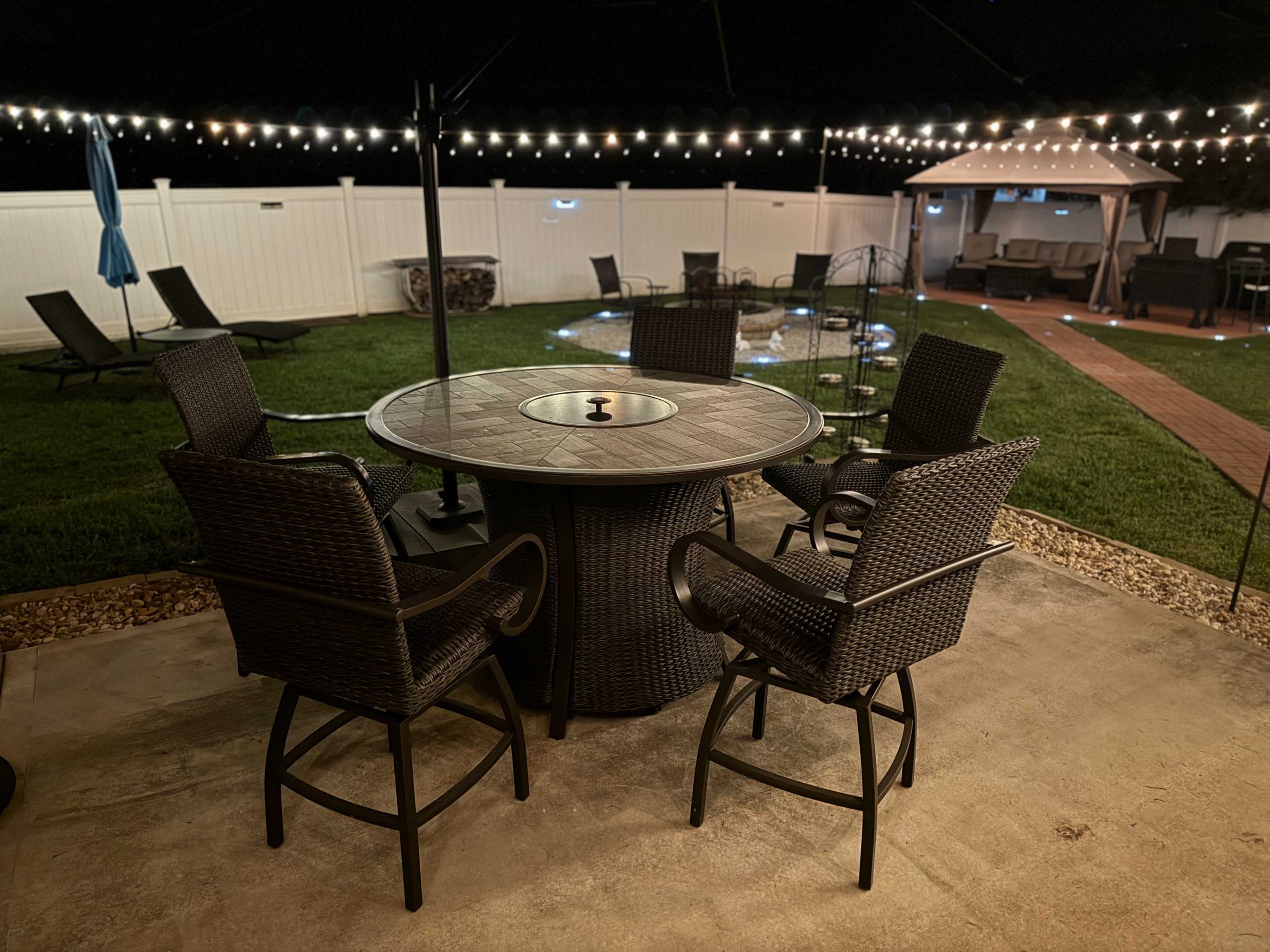 The image shows a patio area with a circular table surrounded by six wicker chairs, illuminated by string lights overhead and featuring a nearby gazebo.