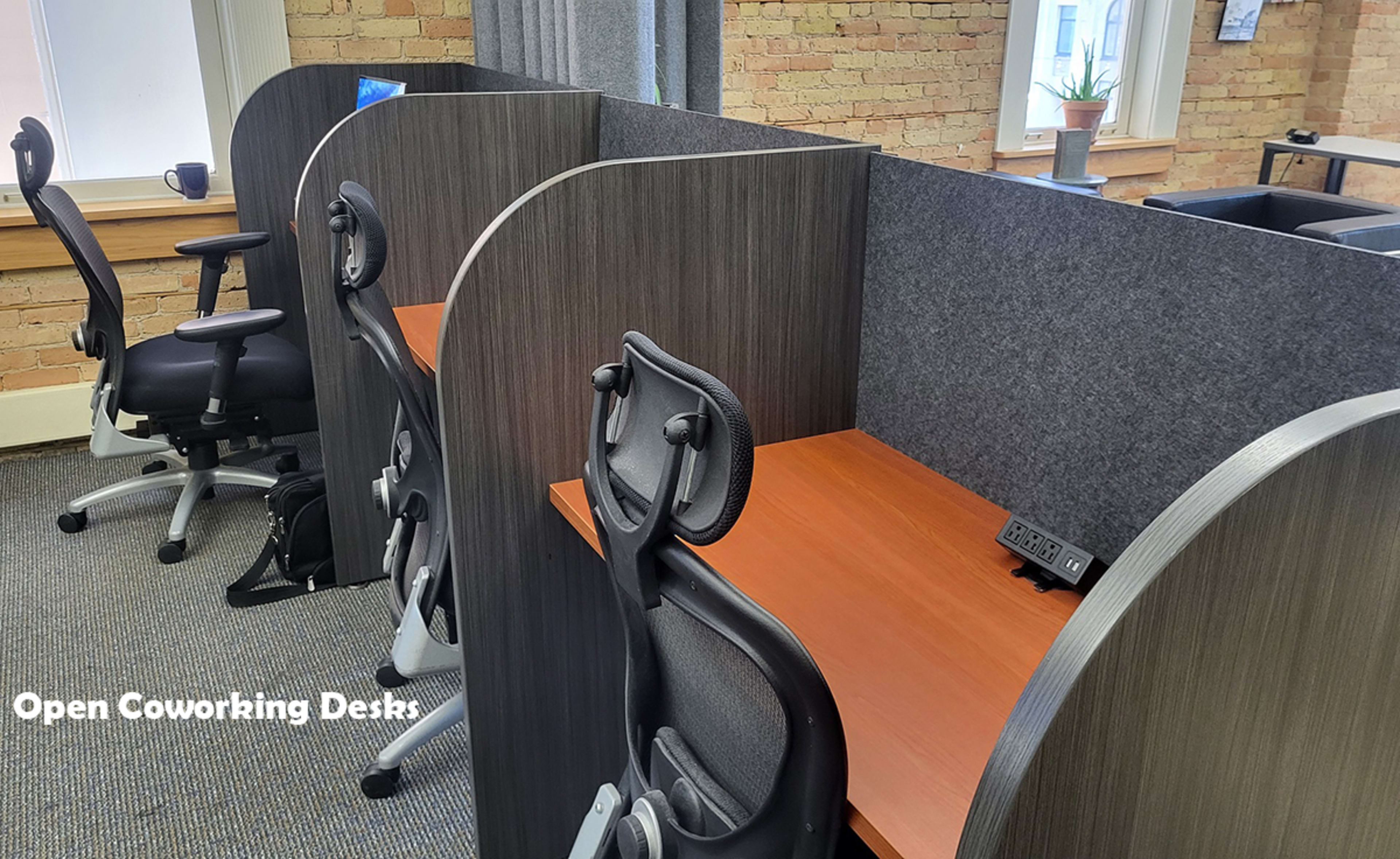 The image shows a row of open coworking desks separated by partitions, with ergonomic chairs positioned at each desk.
