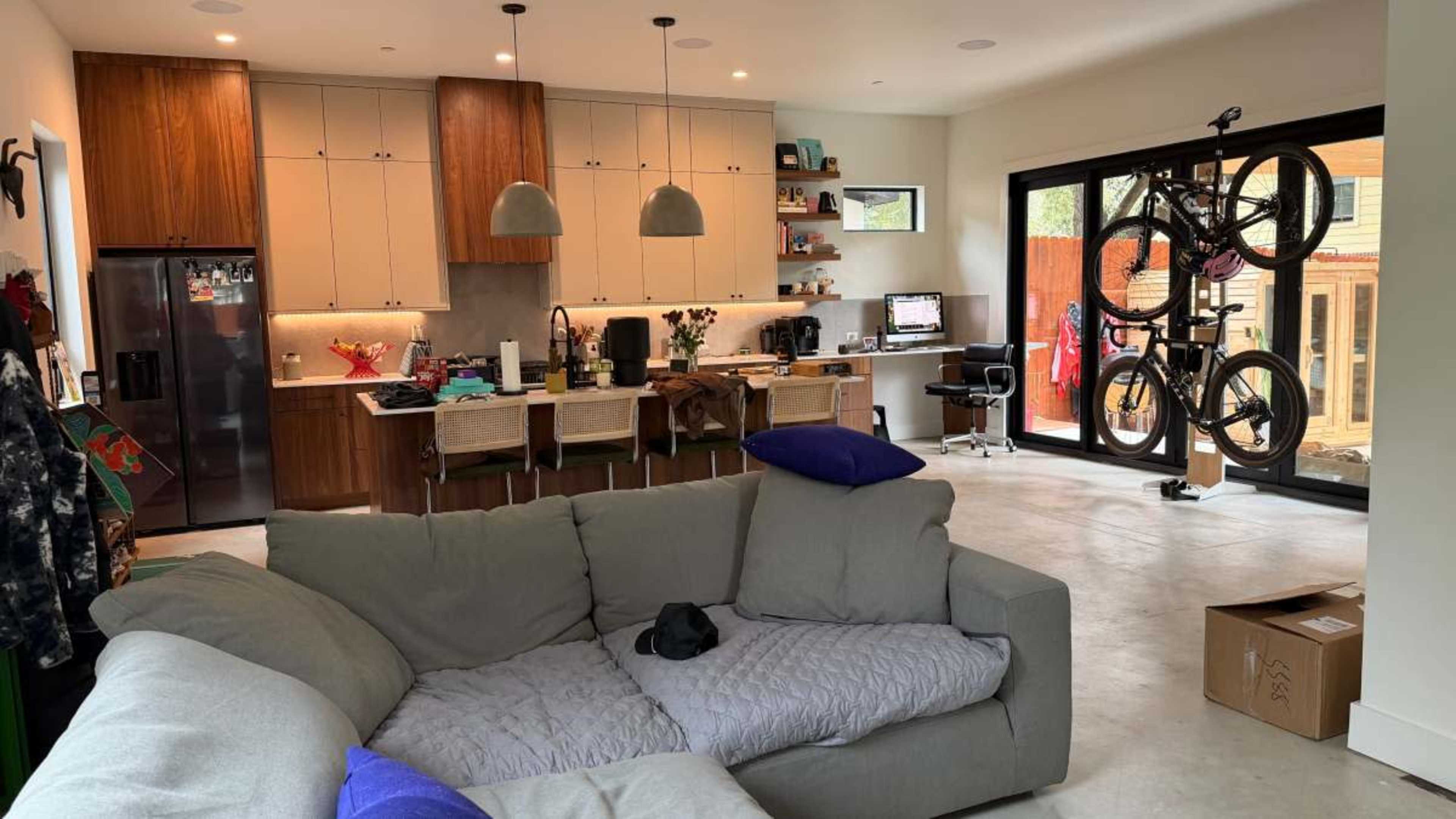 A modern living space features a kitchen with wooden cabinets, a casual seating area with a gray sofa, and bicycles mounted on the wall.