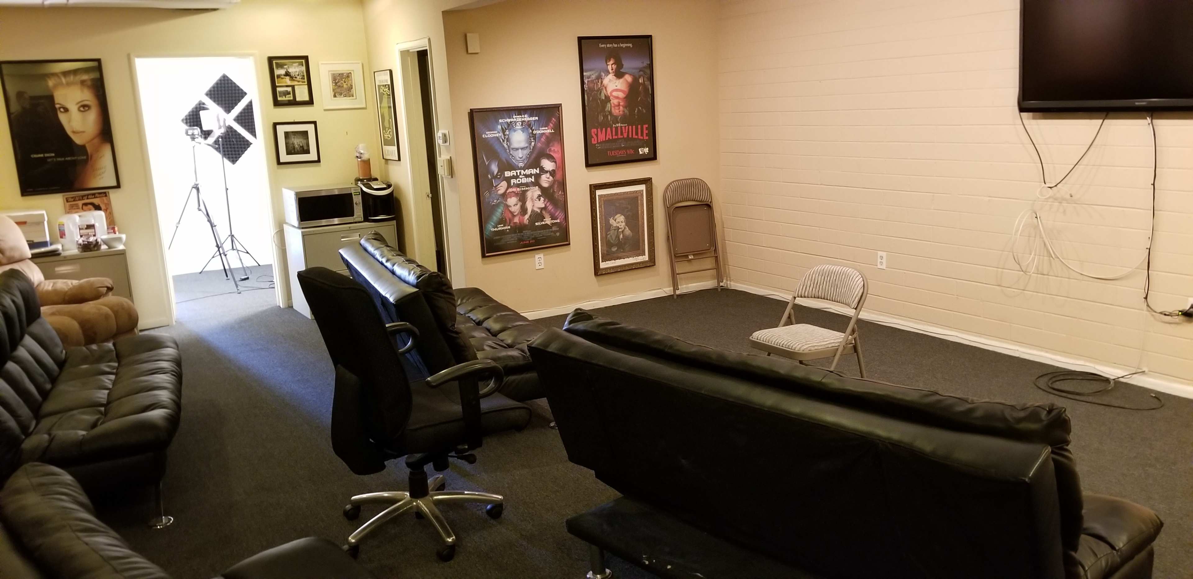 A room with black leather sofas, a single gray chair, several movie posters on the walls, and a television mounted in one corner.