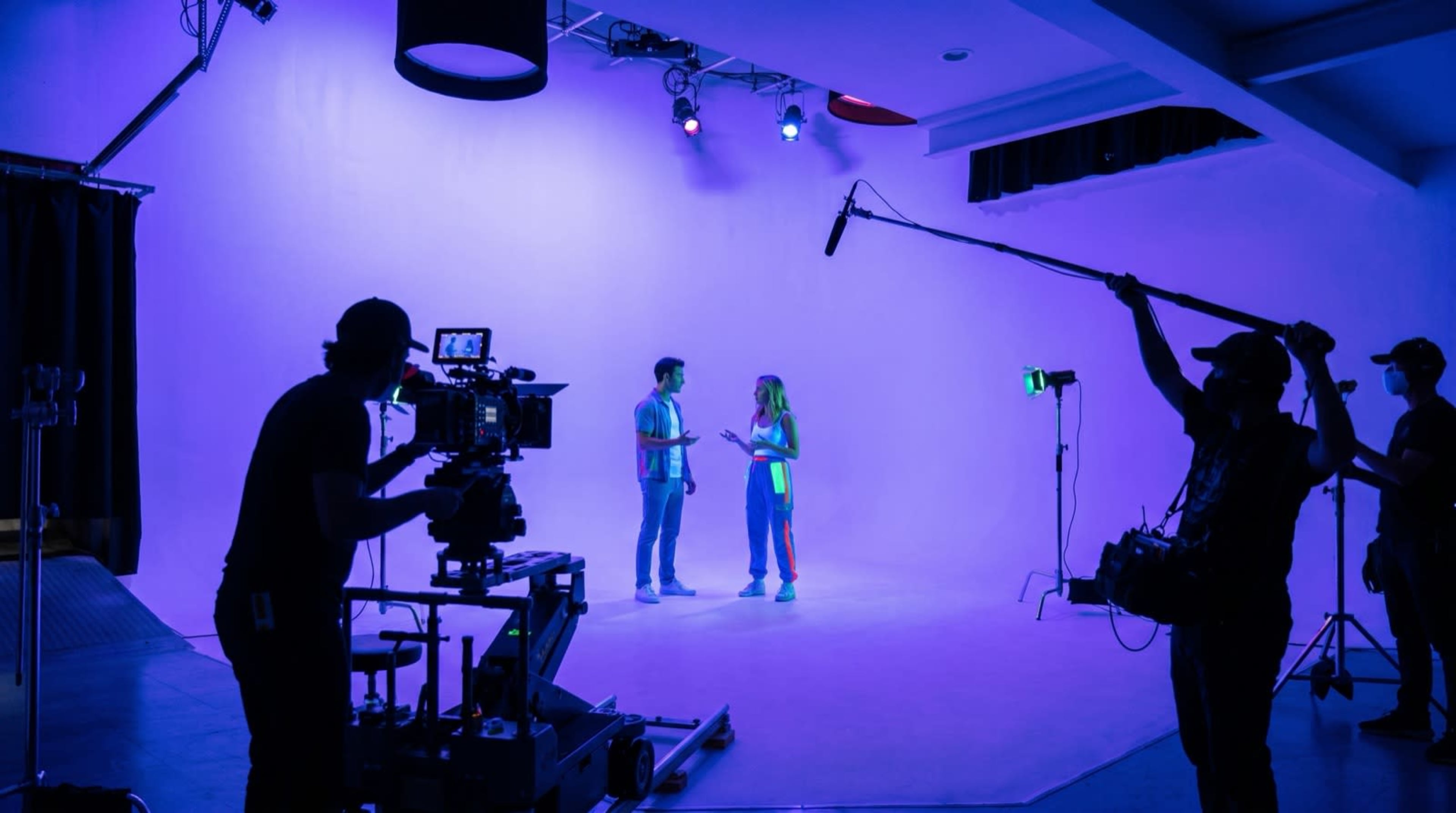 A film crew captures a scene with two actors on a brightly lit, purple backdrop in a studio setting.