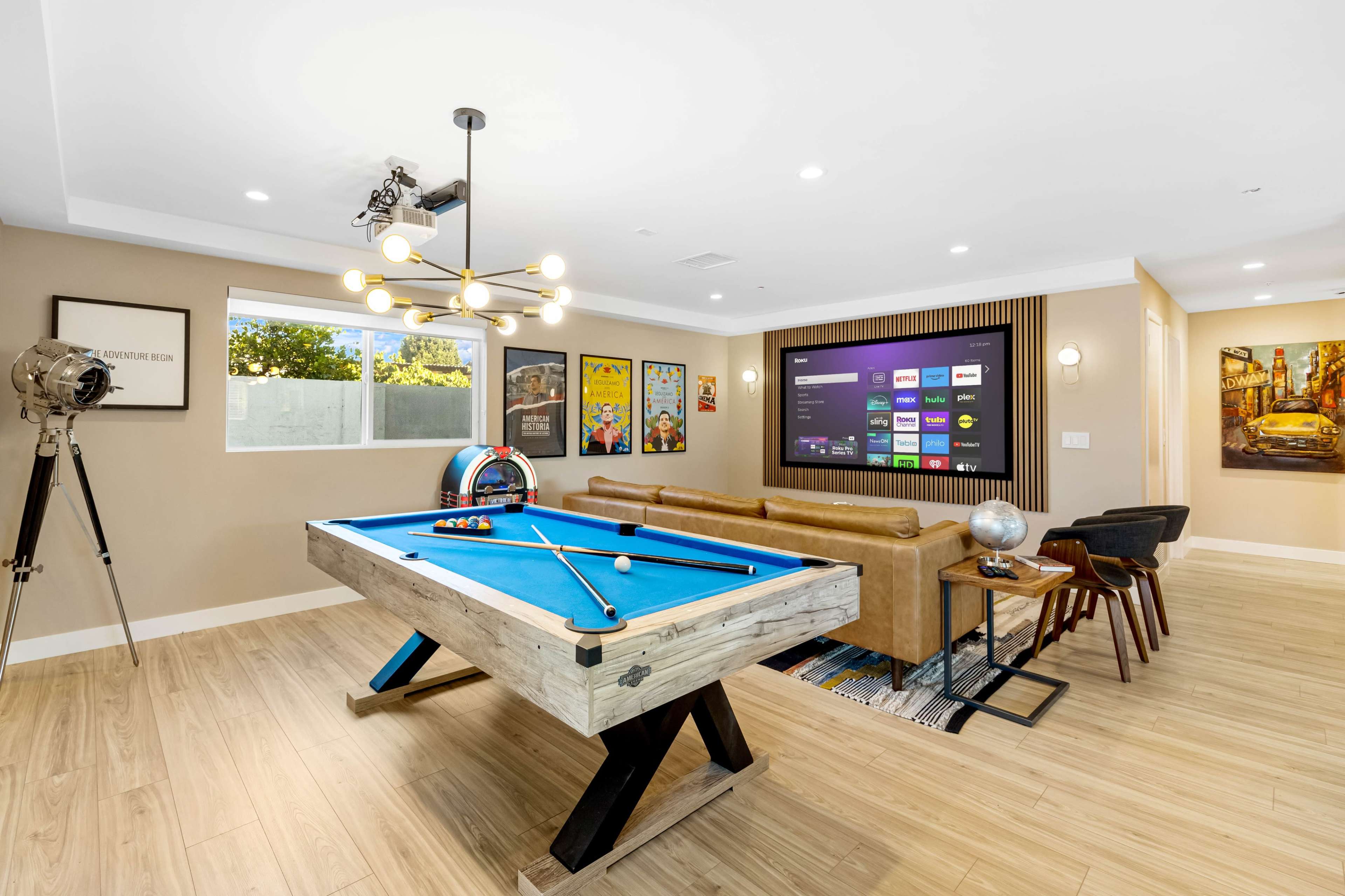 The image shows a modern game room with a blue pool table, a comfortable brown sofa, a large wall-mounted TV, and decorative artwork.