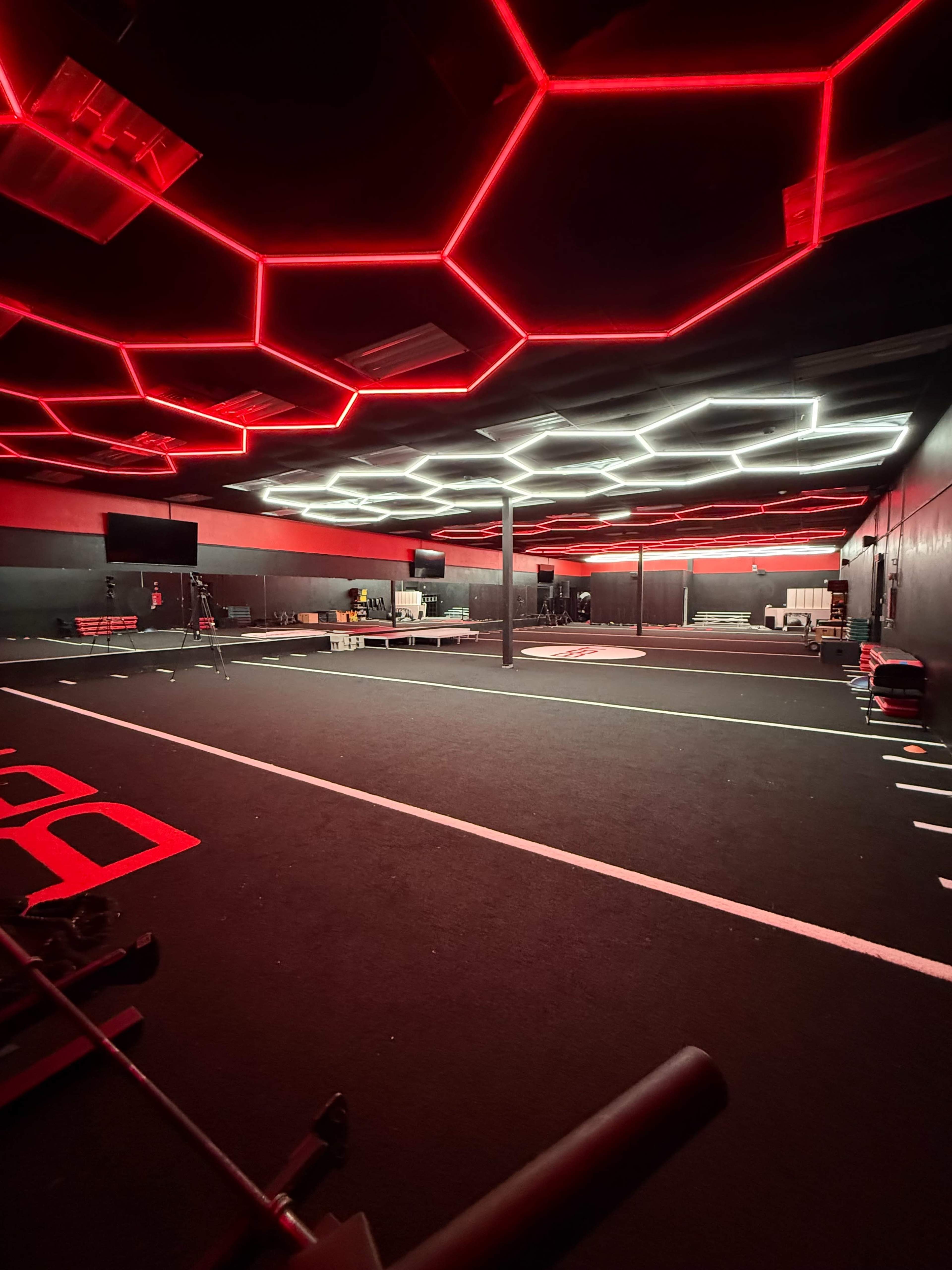 The image shows a dark gym space with a hexagonal ceiling design featuring red and white lights, along with workout equipment arranged on the floor.