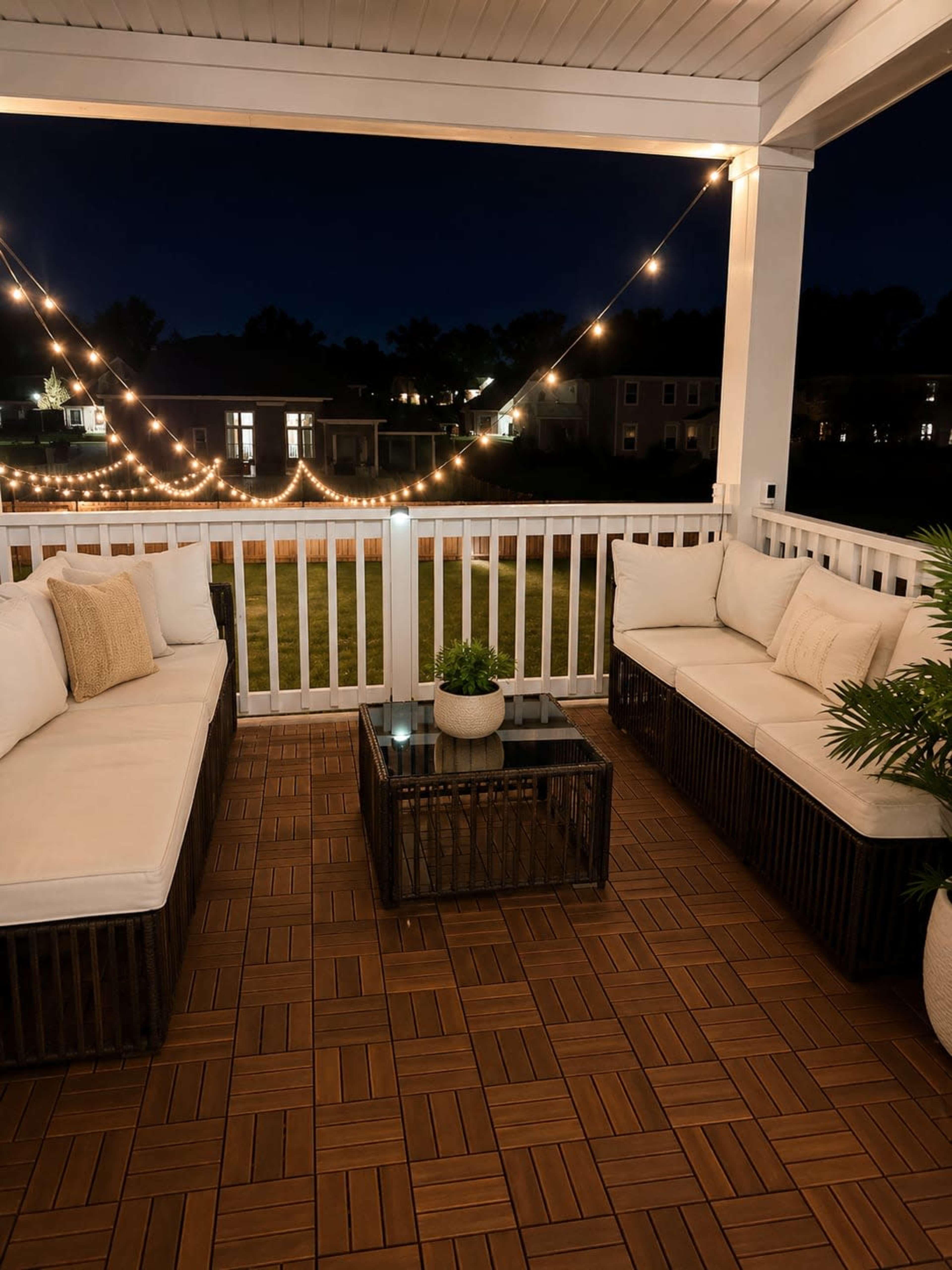 A covered porch features two seating areas with beige cushions, a dark wicker coffee table, and decorative string lights illuminating the night.