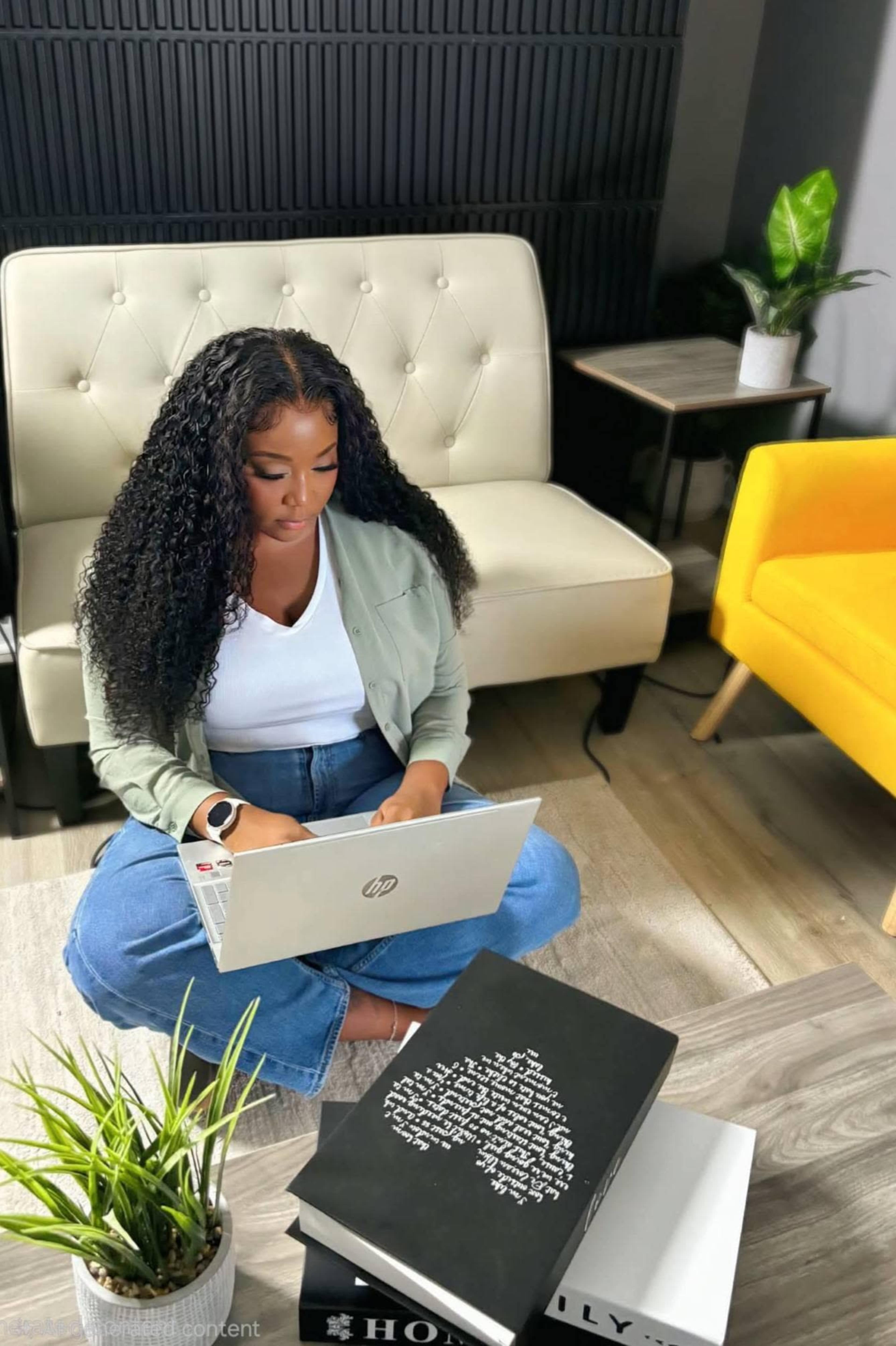 A woman sits on the floor with a laptop, surrounded by books and a plant, in a modern, minimalist room featuring a yellow armchair and a beige sofa.
