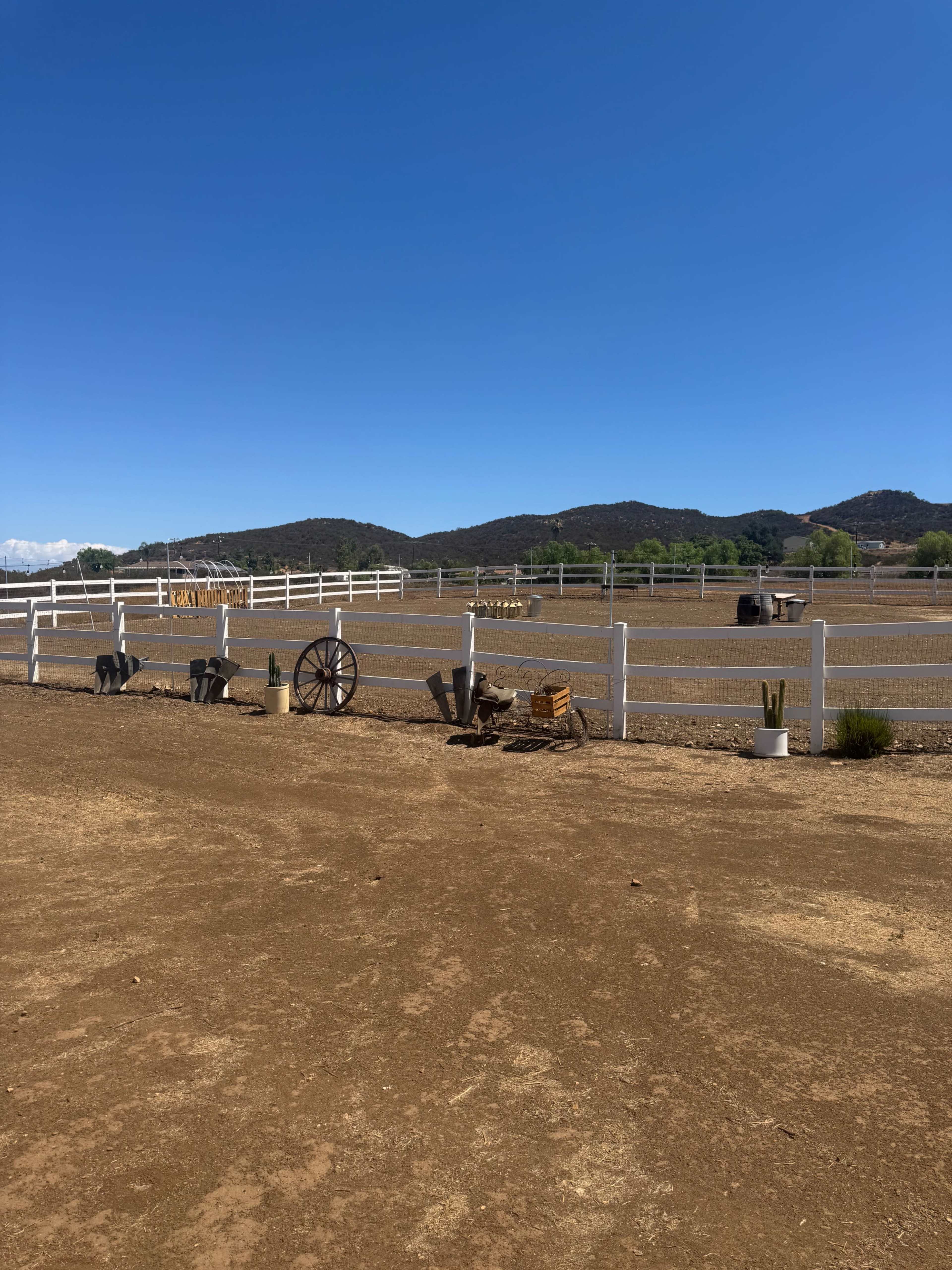 The image shows a fenced dirt area with rustic farm equipment and decorative items in the foreground and rolling hills in the background under a clear blue sky.