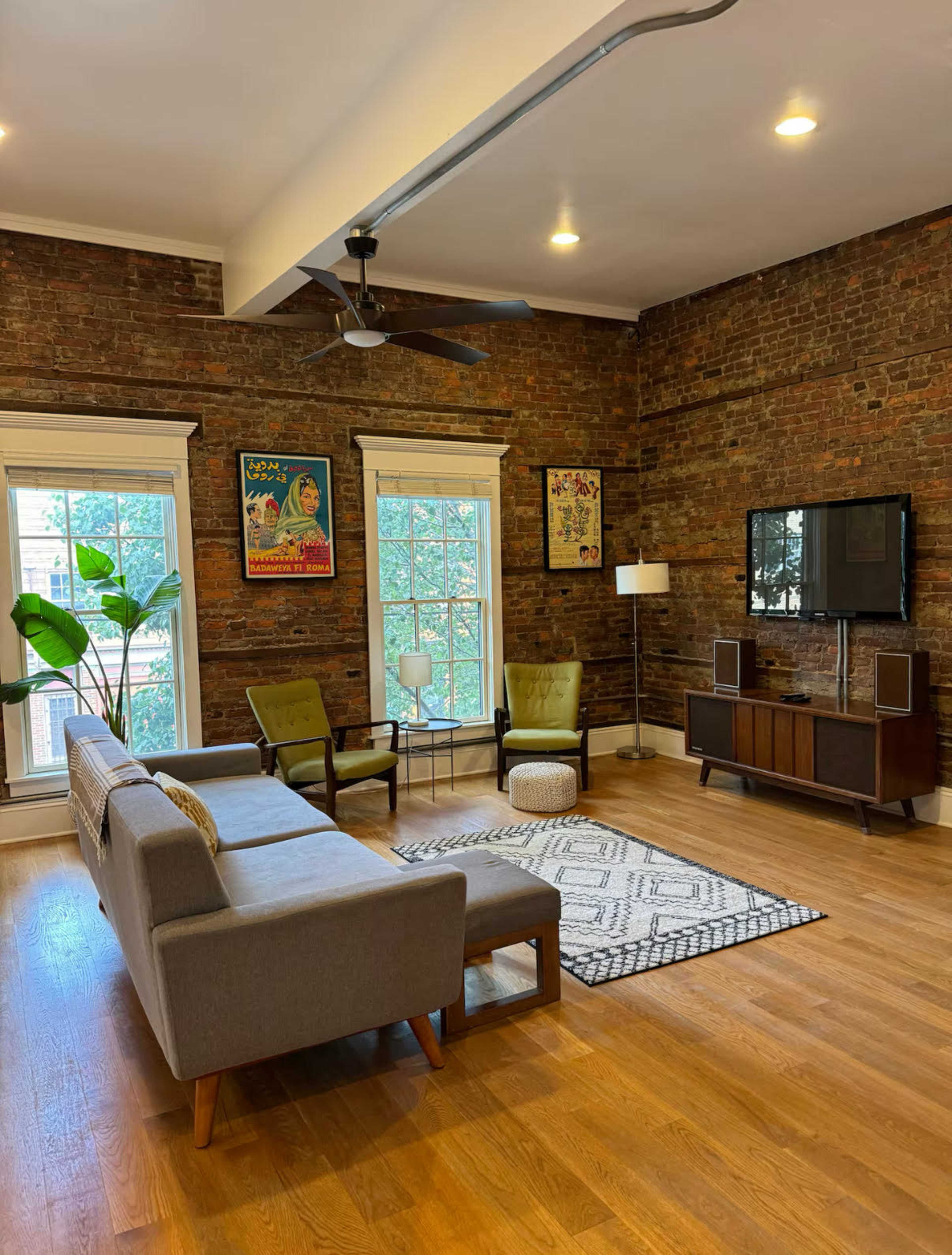 The living room features exposed brick walls, a large window, a sofa, two armchairs, a coffee table, and a television on a media unit.
