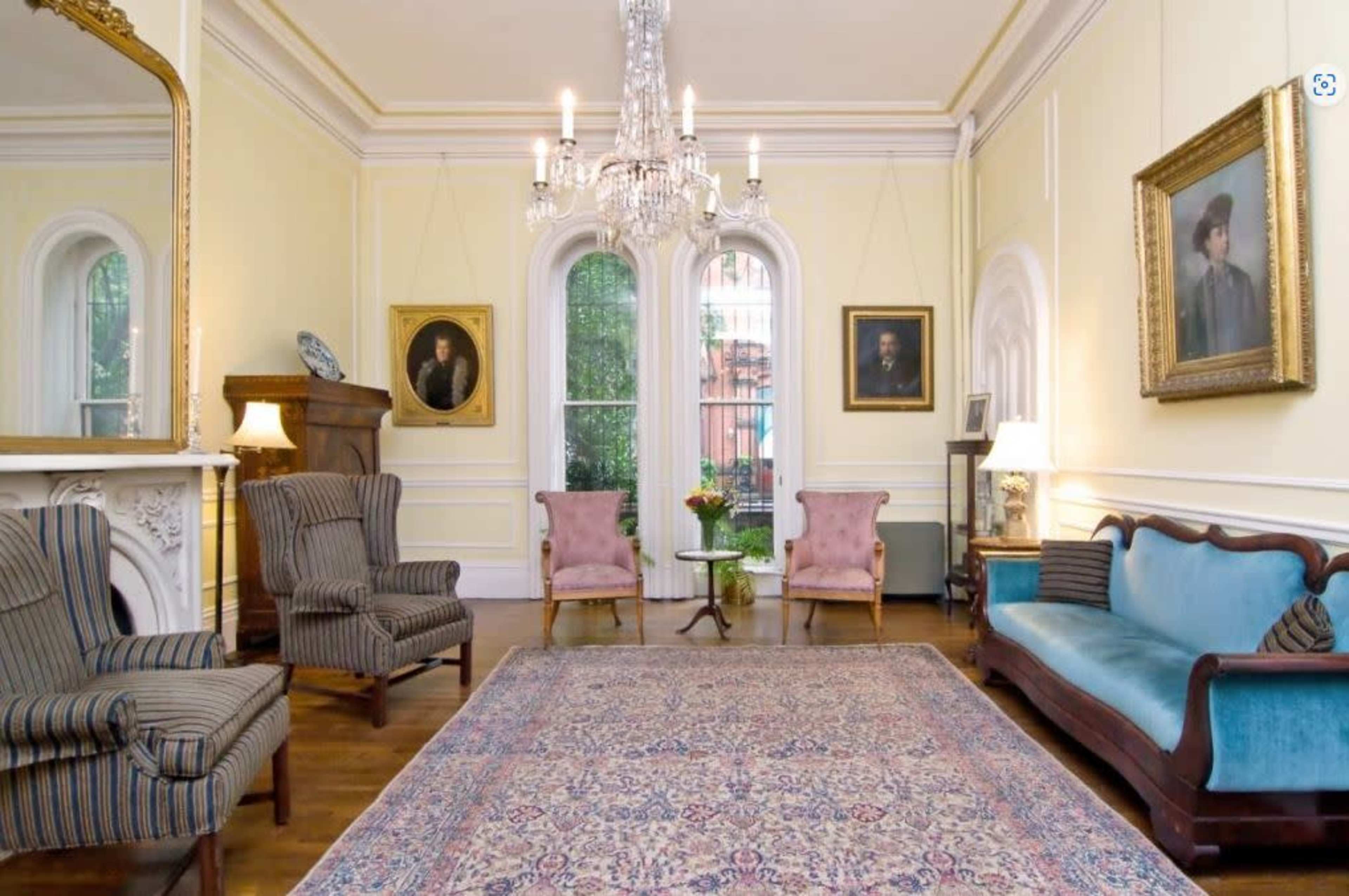 A spacious living room with two striped armchairs, a blue sofa, a chandelier, and various framed portraits on the yellow walls.