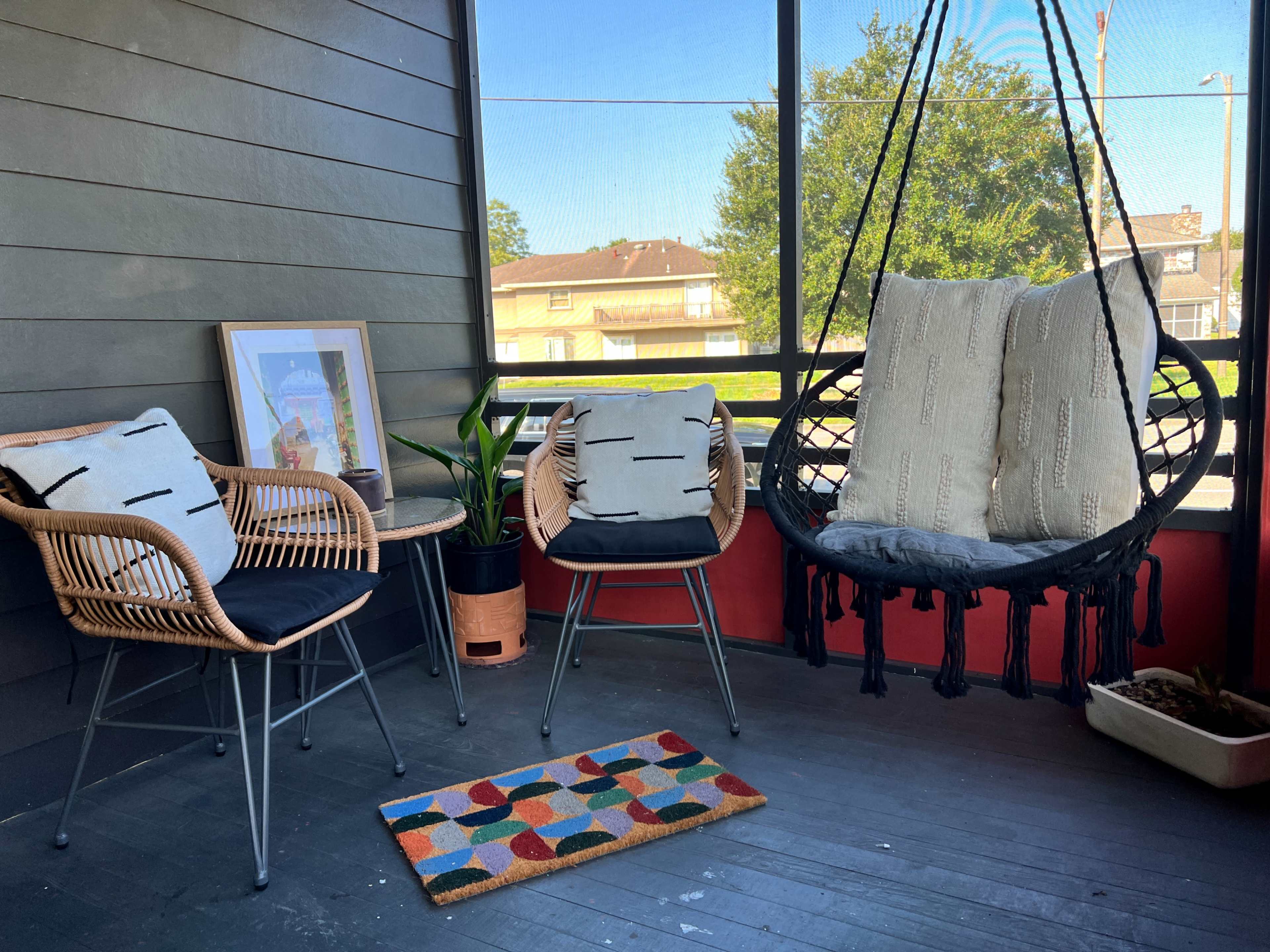 The image shows a screened-in porch with two chairs, a hanging chair, a potted plant, and a colorful rug.