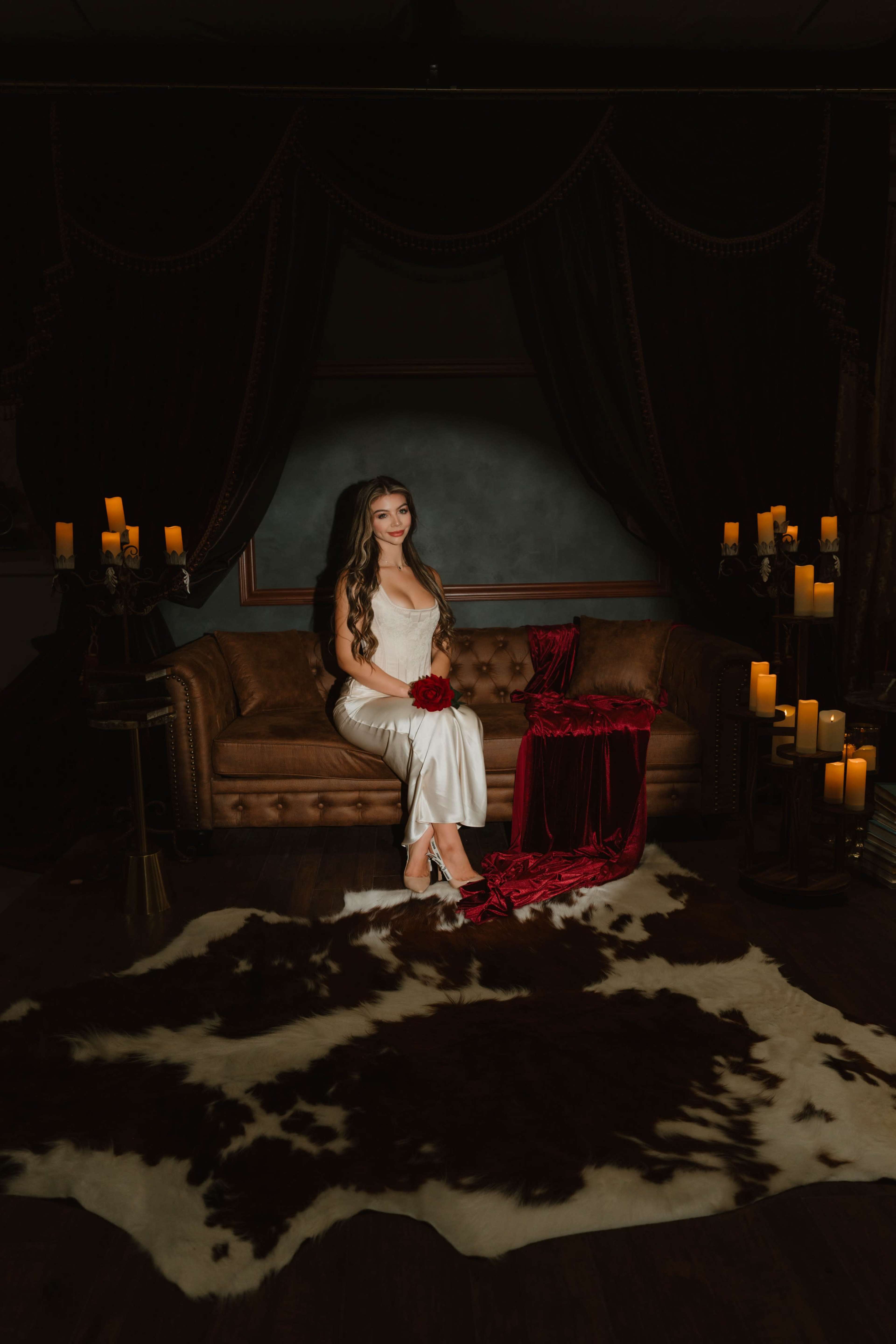 A woman sits on a vintage couch draped in a red cloth, surrounded by candles and a cowhide rug.