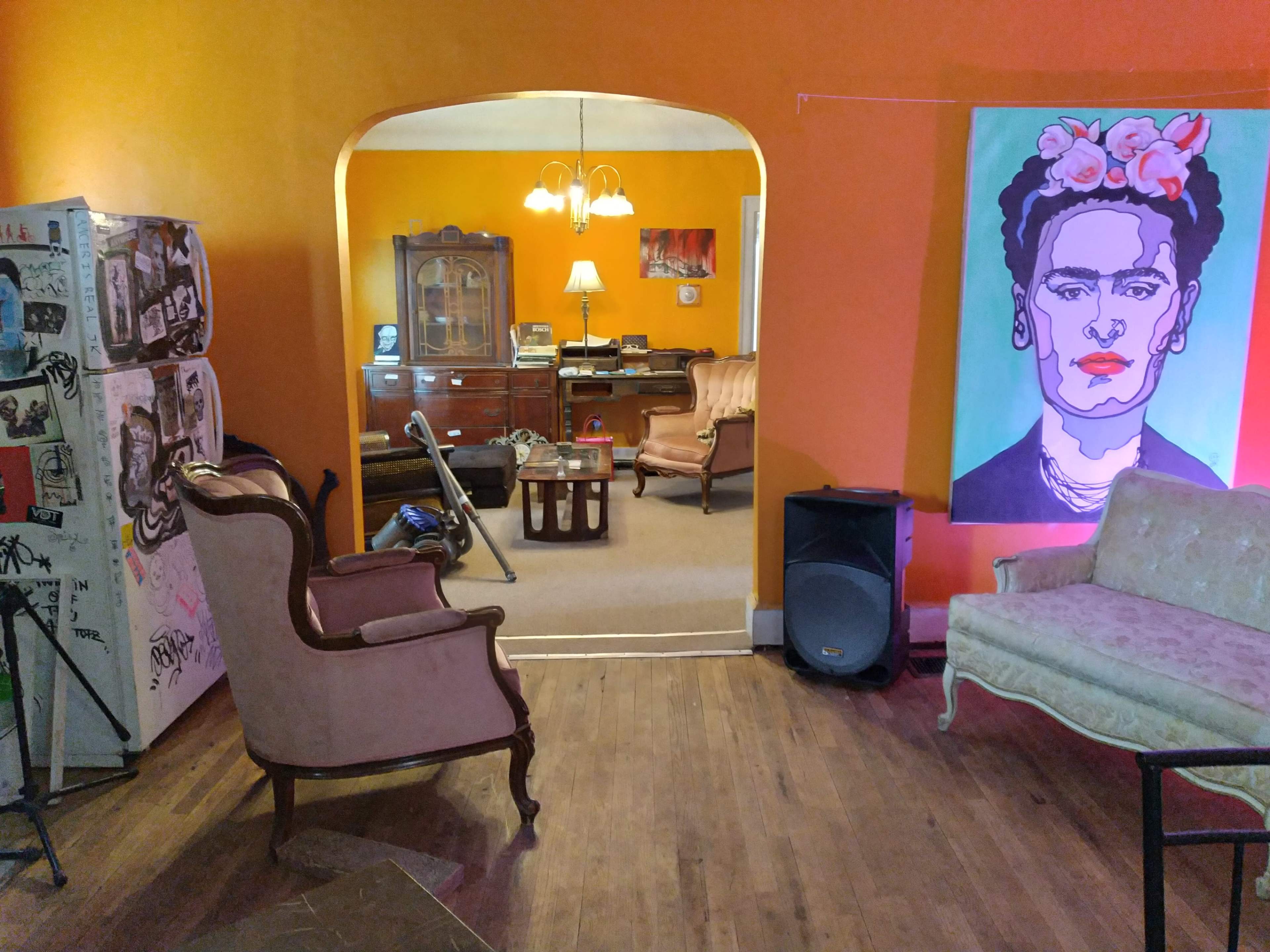 The image shows a brightly painted orange room with a vintage decor featuring two armchairs, a small coffee table, a refrigerator covered in stickers, and a colorful portrait of Frida Kahlo on the wall.
