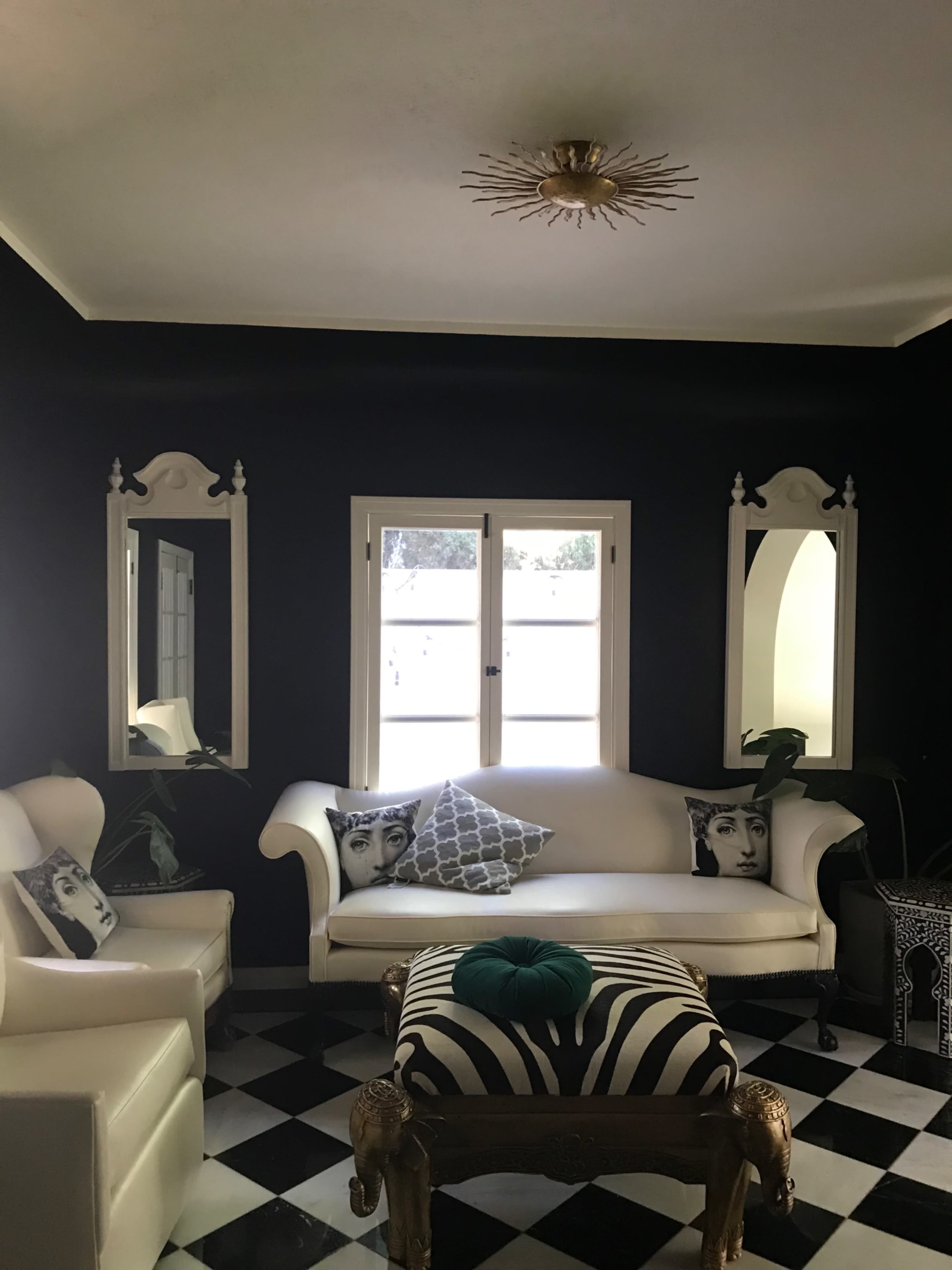 The room features a black wall, a white couch with decorative pillows, and a checked black and white floor, complemented by mirrors and a brass light fixture.
