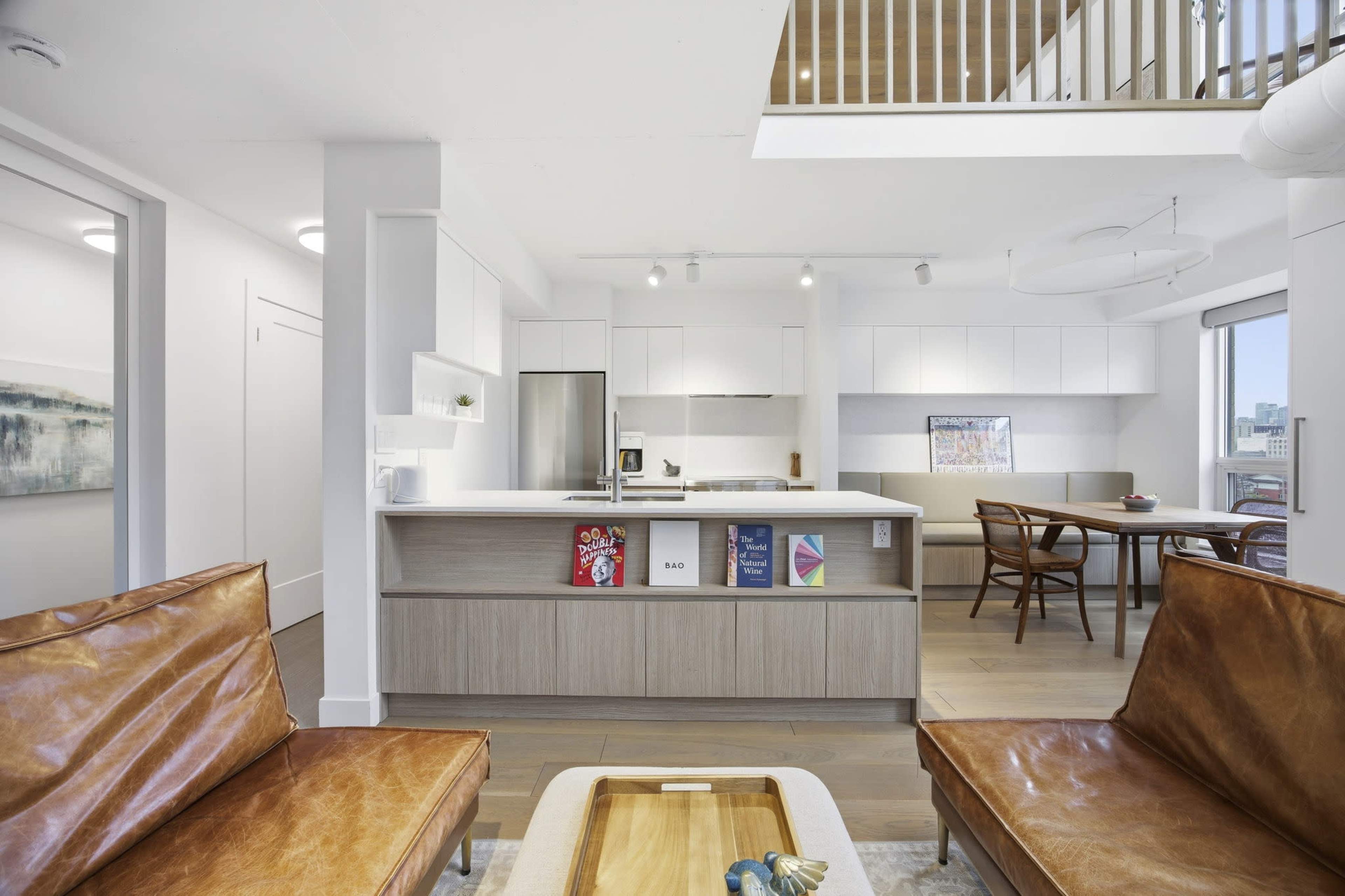 The image shows a modern, open-concept living space featuring a kitchen, dining area, and lounge with two brown leather chairs and a wood table.