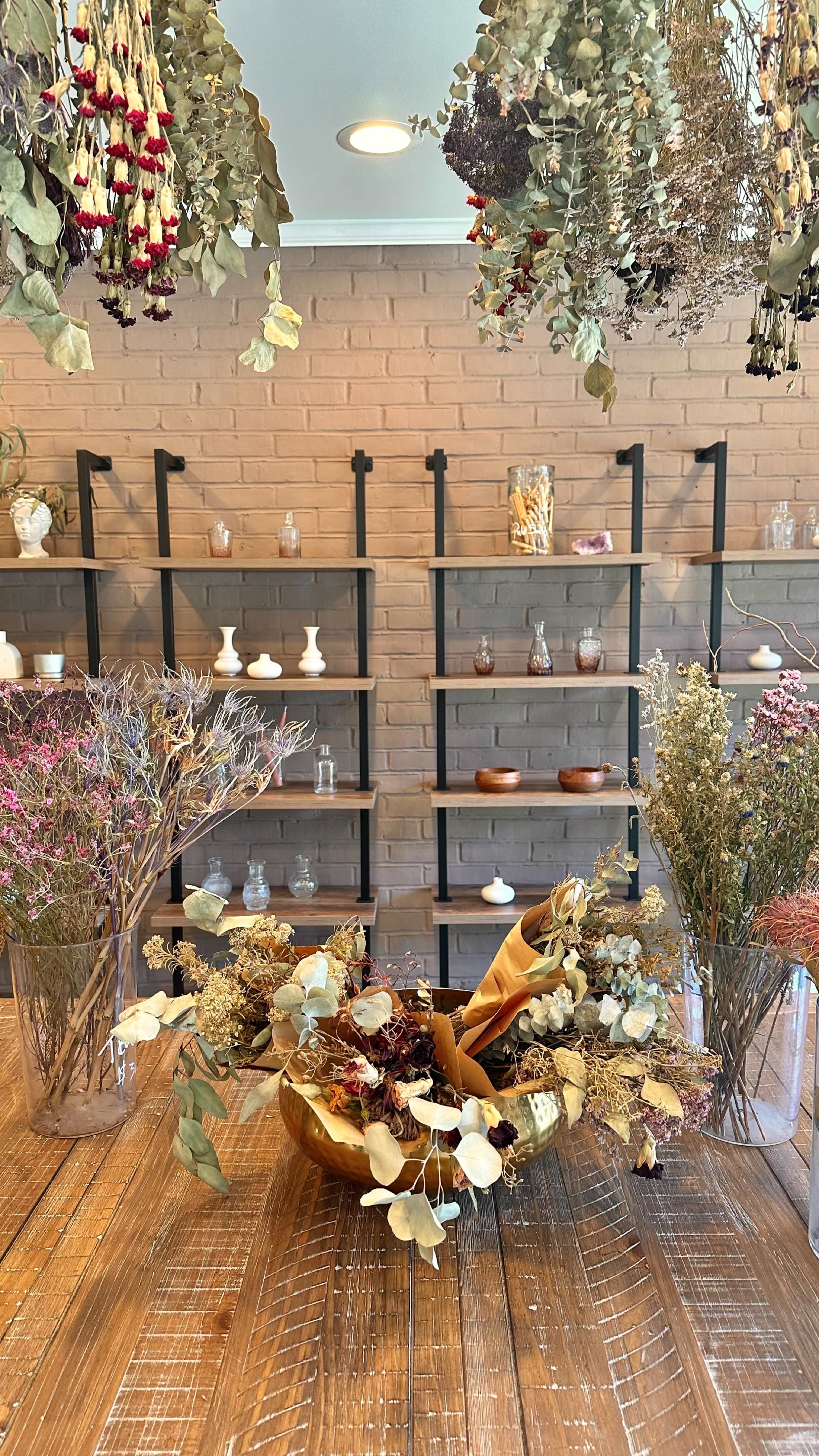 A rustic interior with wooden tables, metal shelving displaying various glass jars and decorative items, and an arrangement of dried flowers and plants in a wooden bowl.