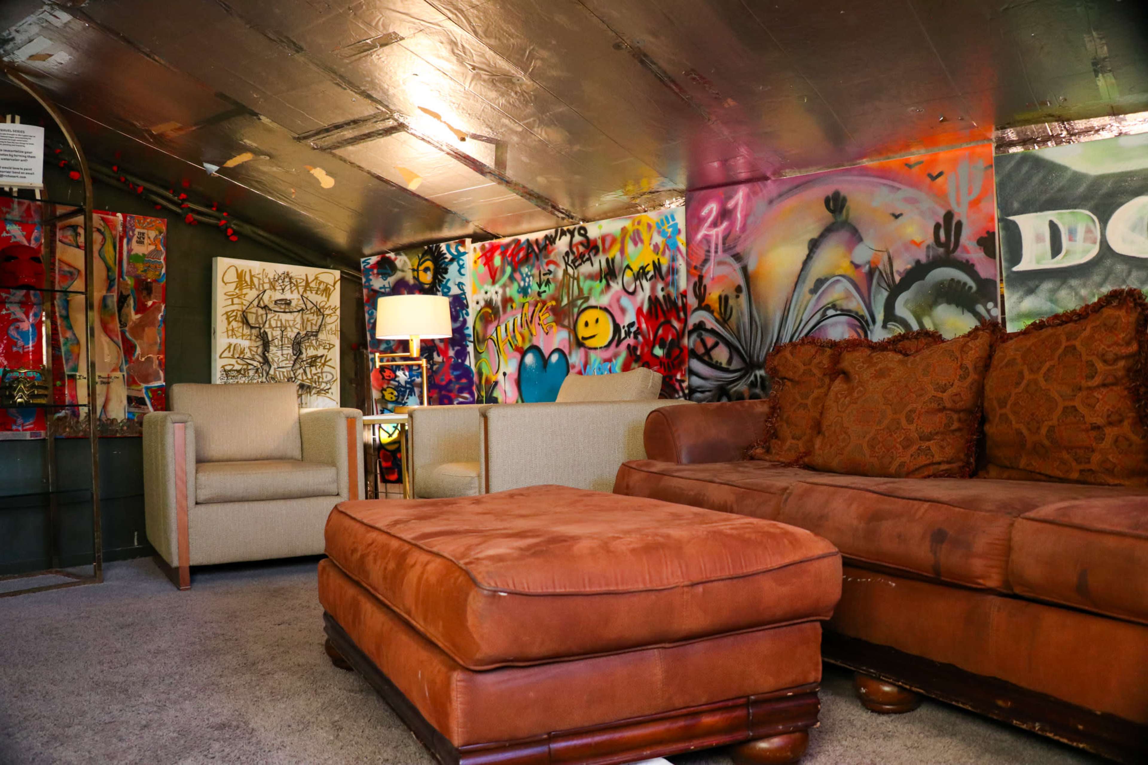 A small attic-style lounge area with colorful graffiti on the walls, featuring a brown sofa, an ottoman, and two light-colored armchairs.