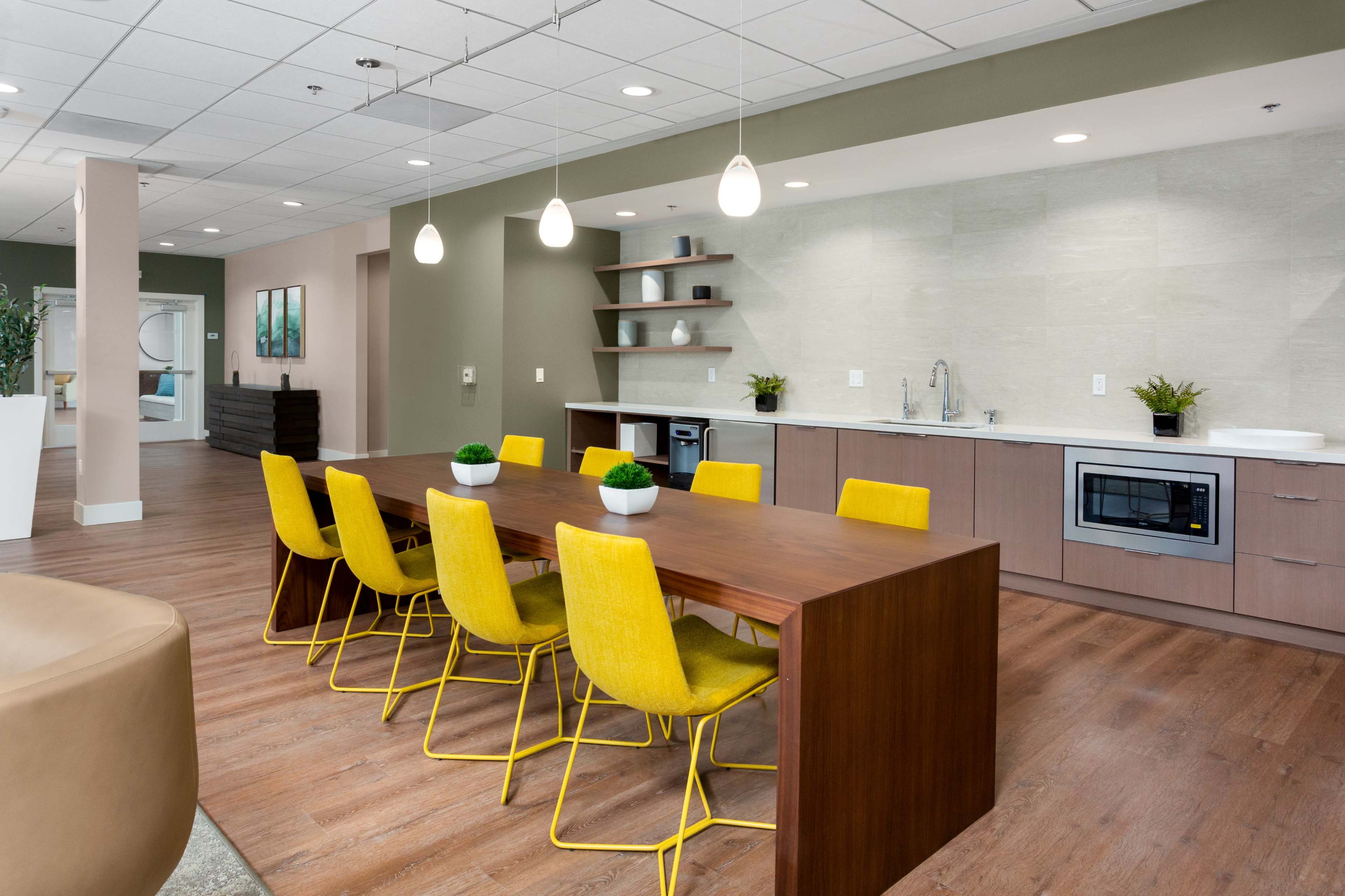 A modern dining area features a long wooden table surrounded by yellow chairs, with a kitchenette and shelves in the background.
