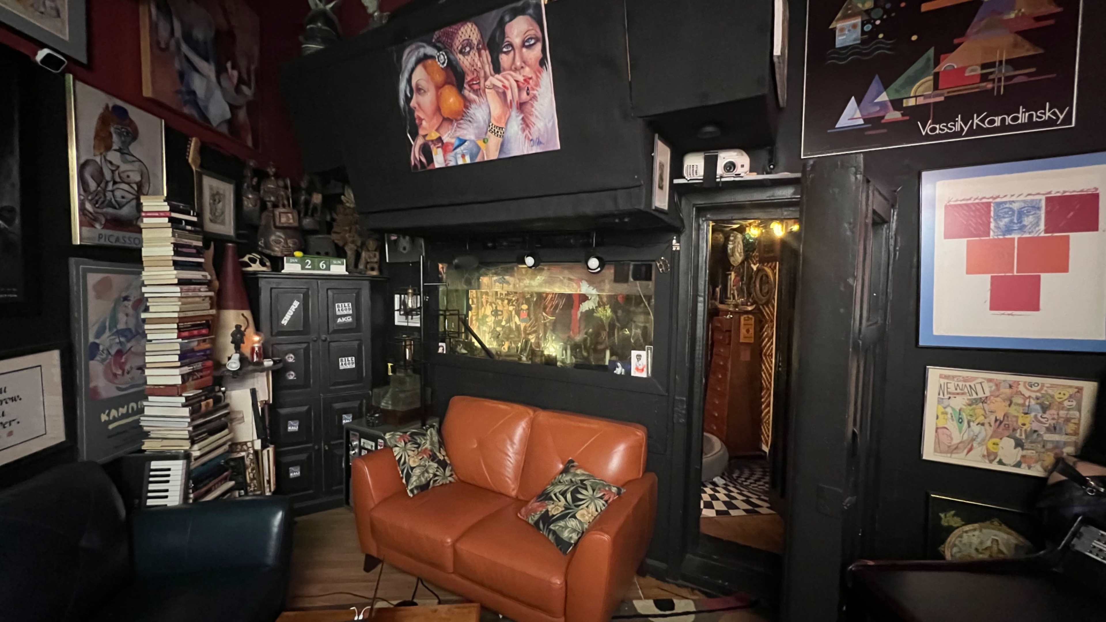A cozy room with an orange sofa, shelves filled with books, and various artworks hanging on the dark walls.