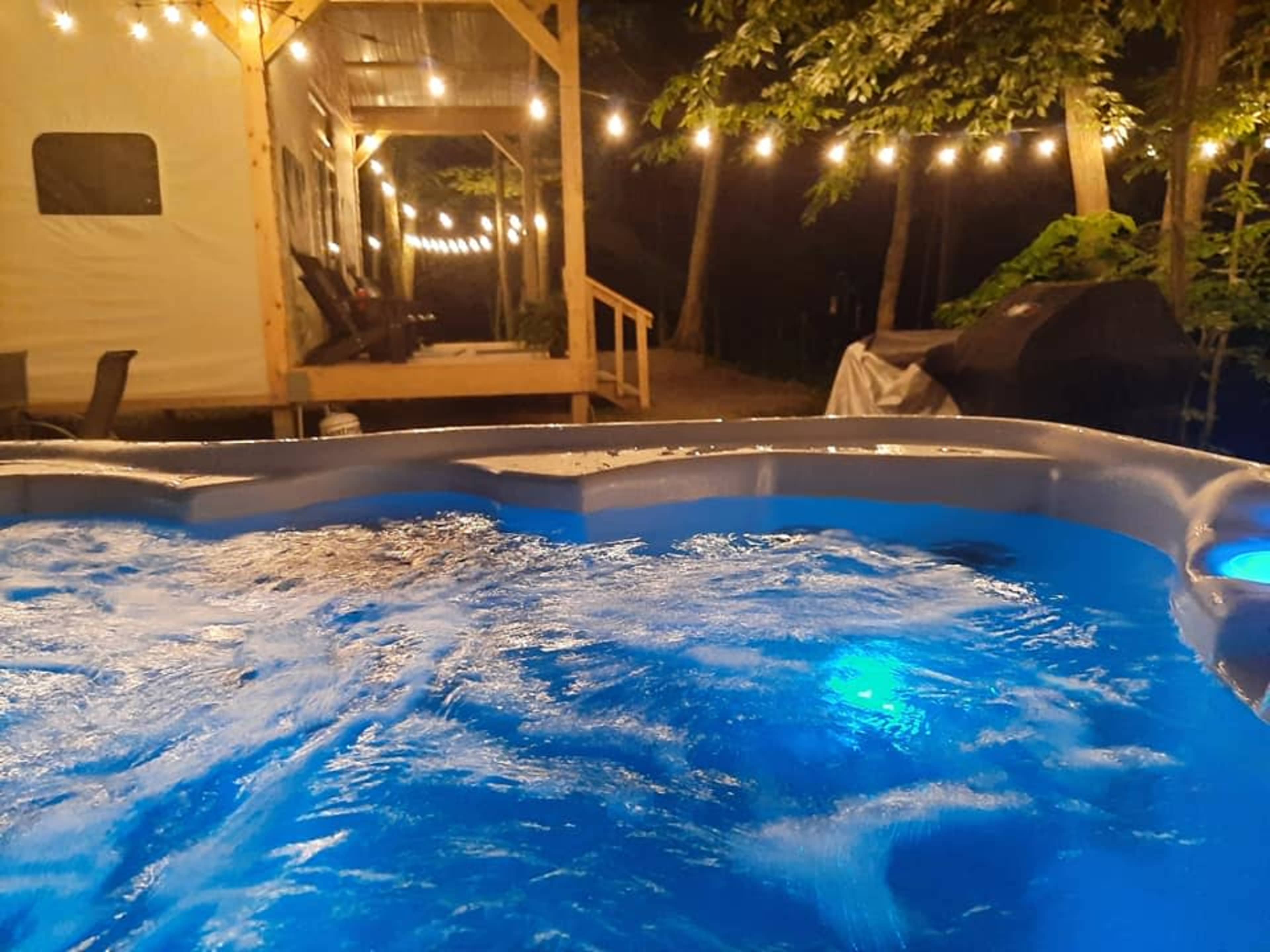 A hot tub illuminated with blue lights, situated near a wooden deck adorned with string lights in a wooded outdoor area at night.