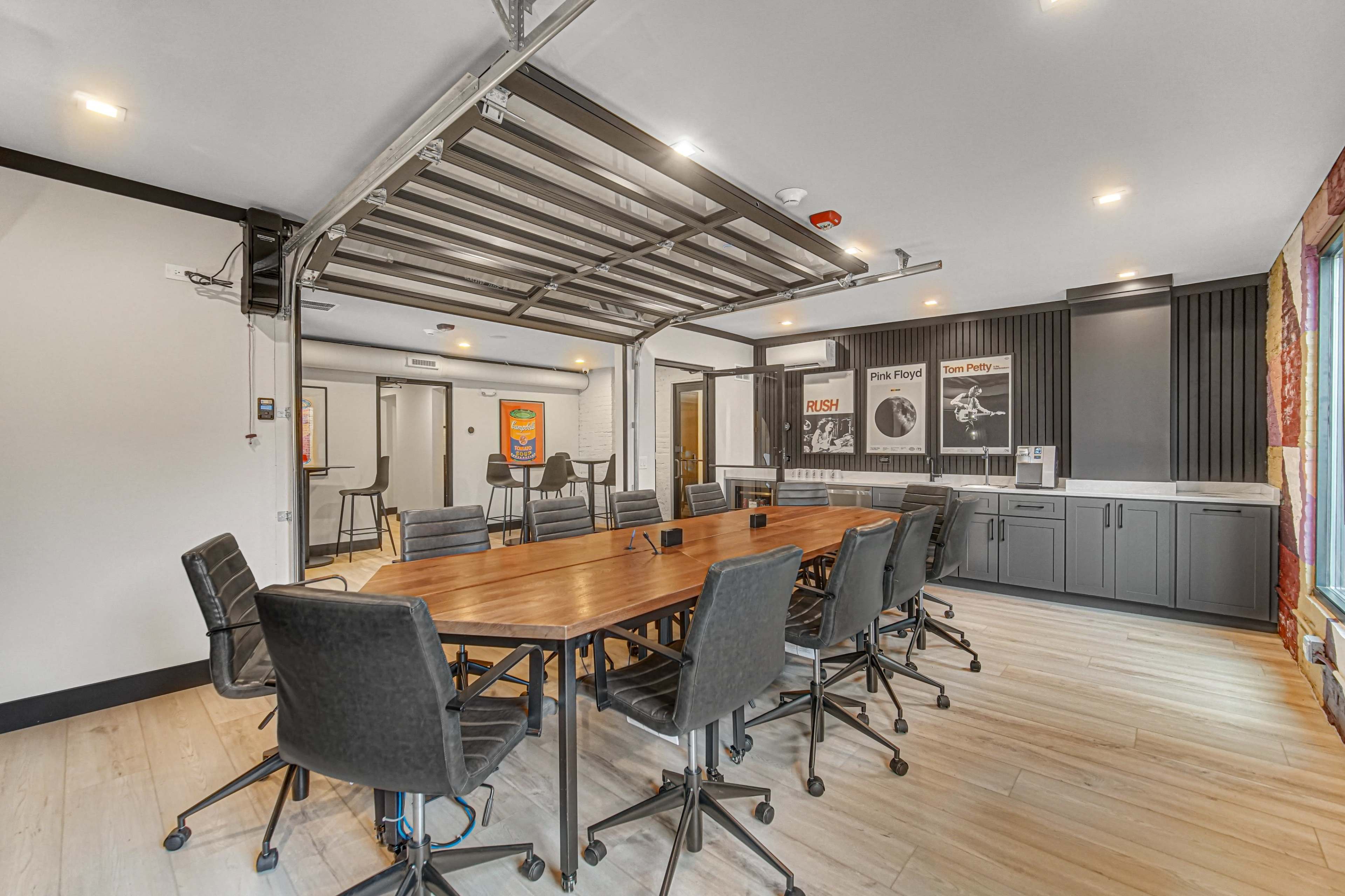 A modern conference room features a large wooden table surrounded by black rolling chairs, with a retractable ceiling and framed posters on the walls.
