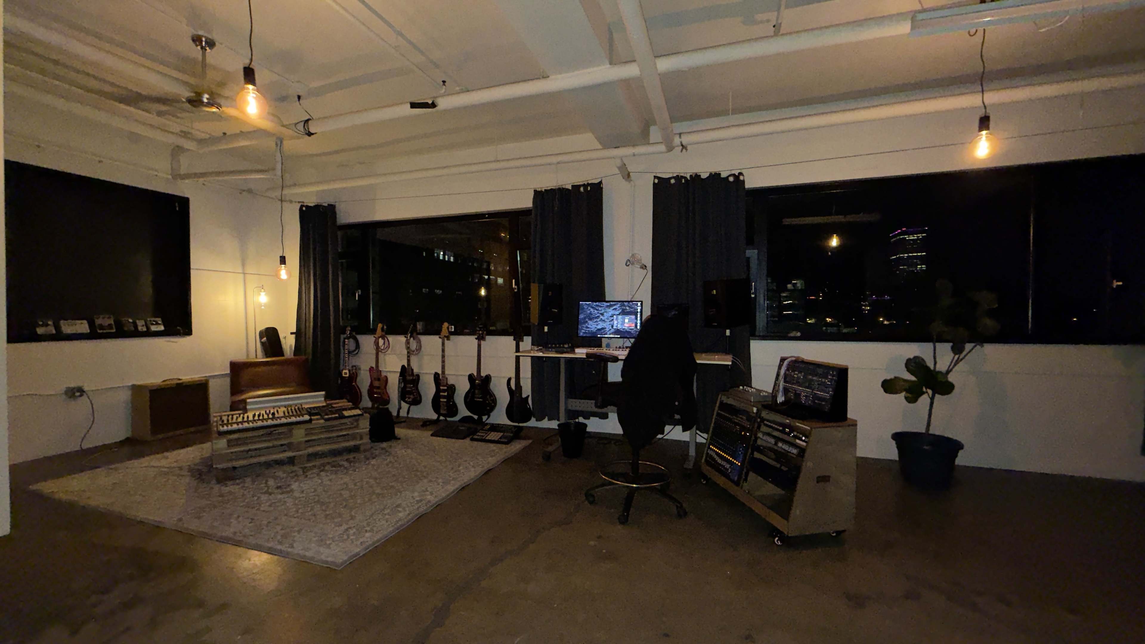 The image shows a music studio with instruments, a desk with equipment, and large windows displaying a nighttime city skyline.