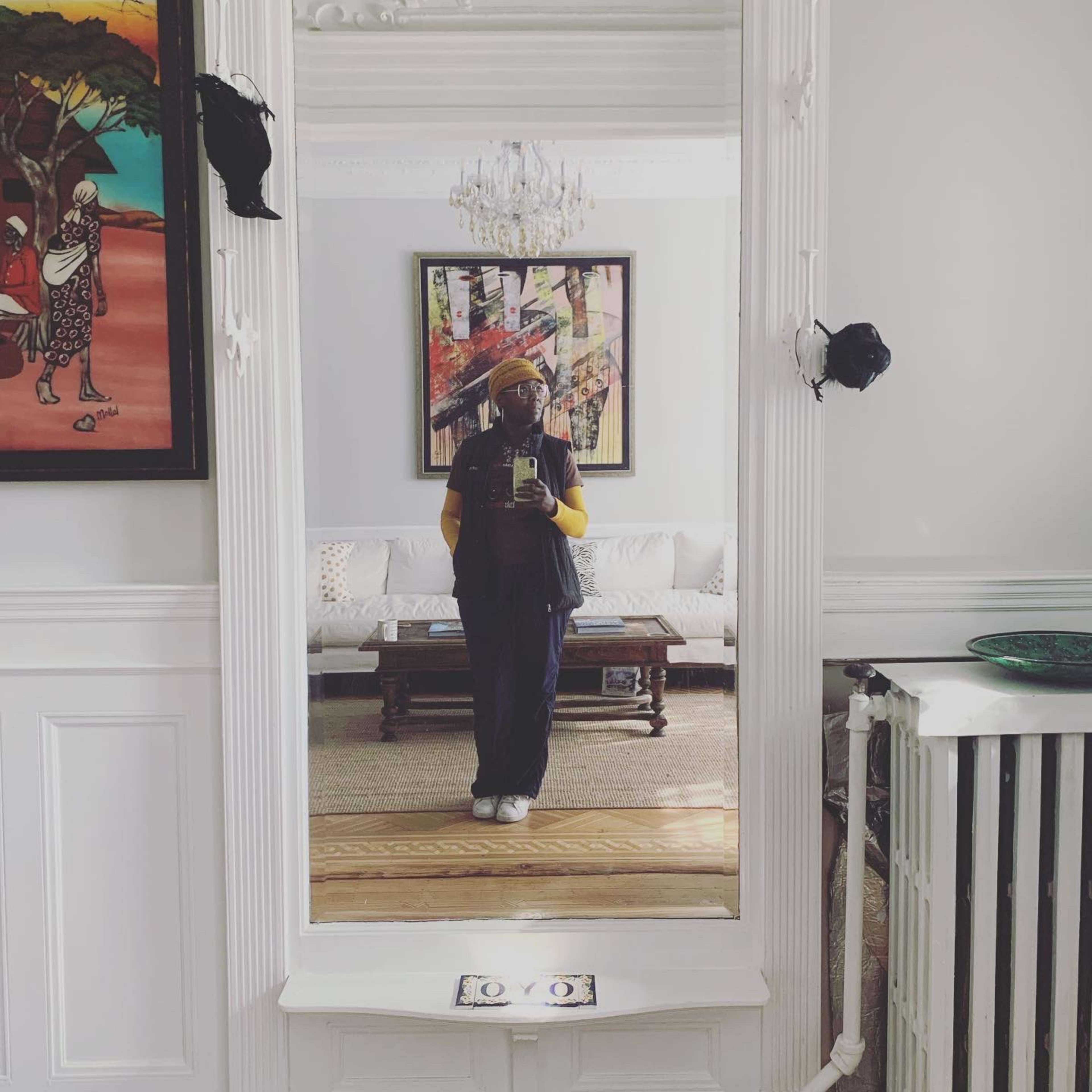 A person stands in front of a large mirror in a bright room decorated with a chandelier and artwork, with a sofa and rug visible in the background.