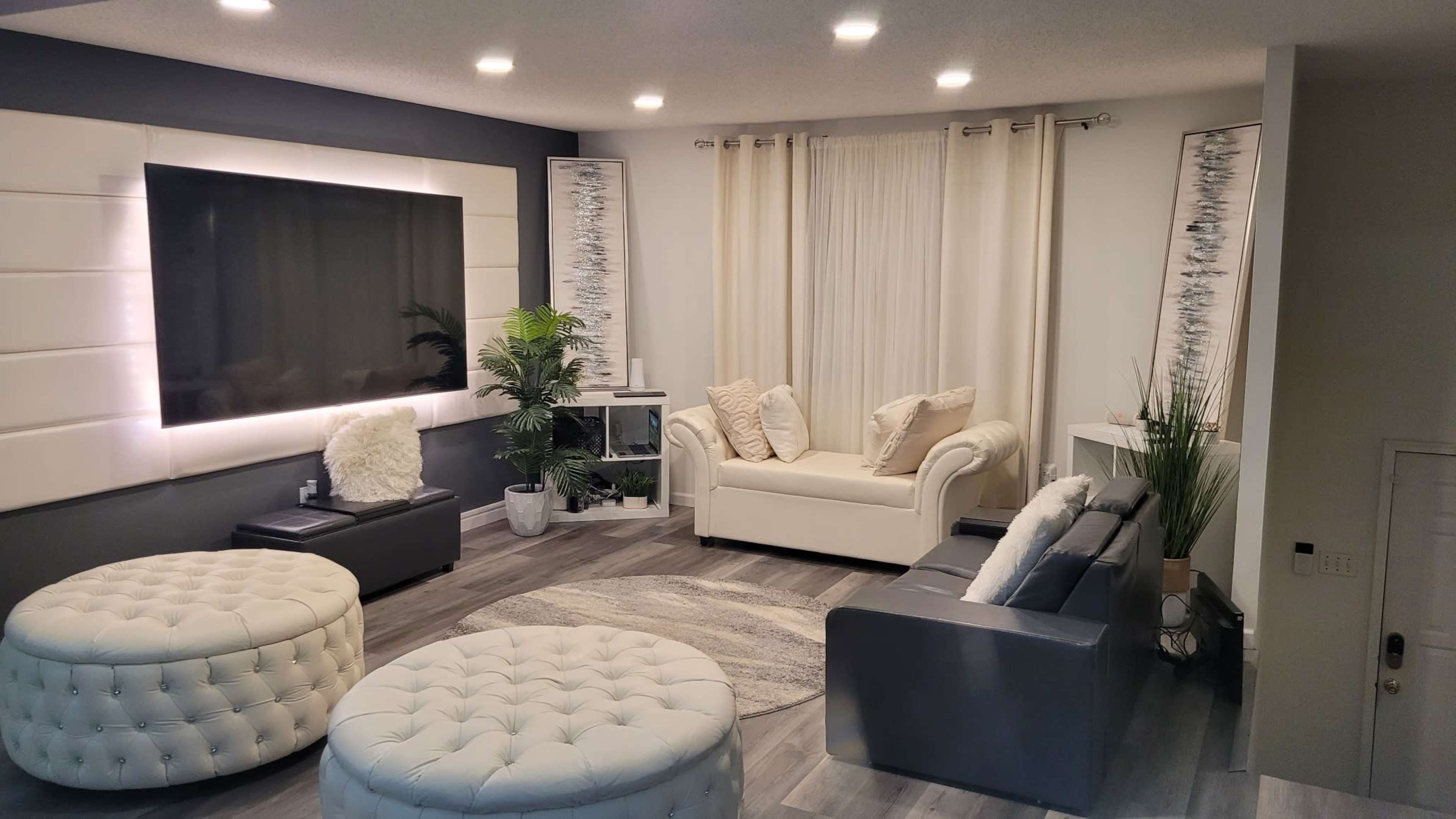 A modern living room with white sofas, round ottomans, and a large television against a feature wall, complemented by plants and curtains.