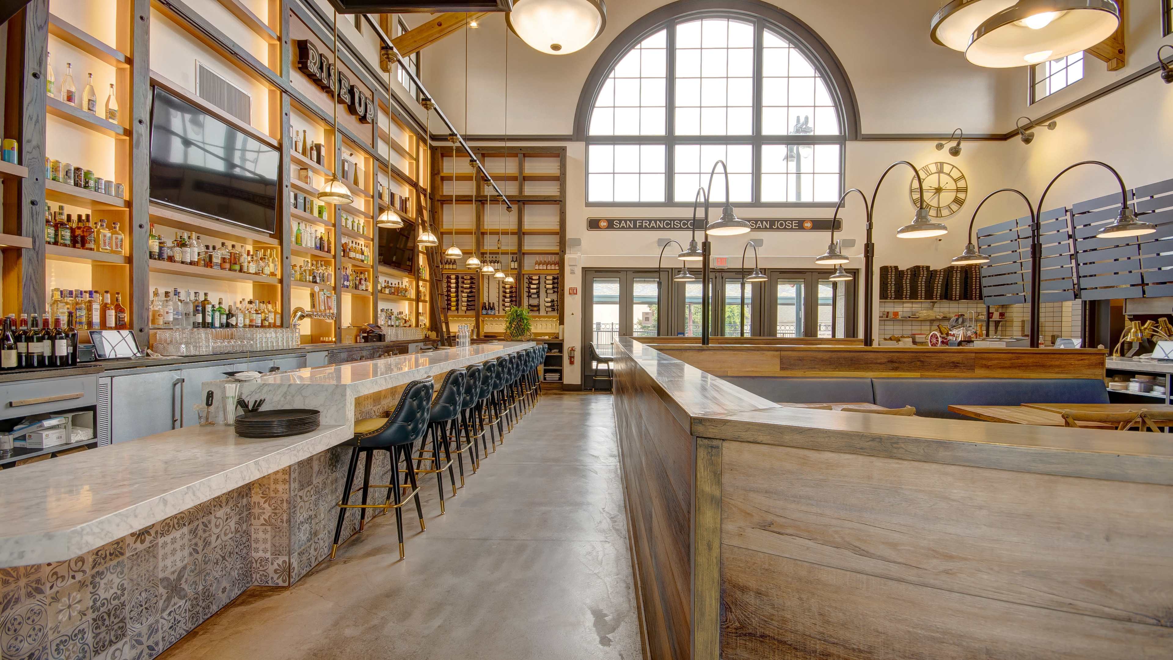 A modern restaurant interior with a long bar, high stools, and large windows allowing natural light to illuminate the space.