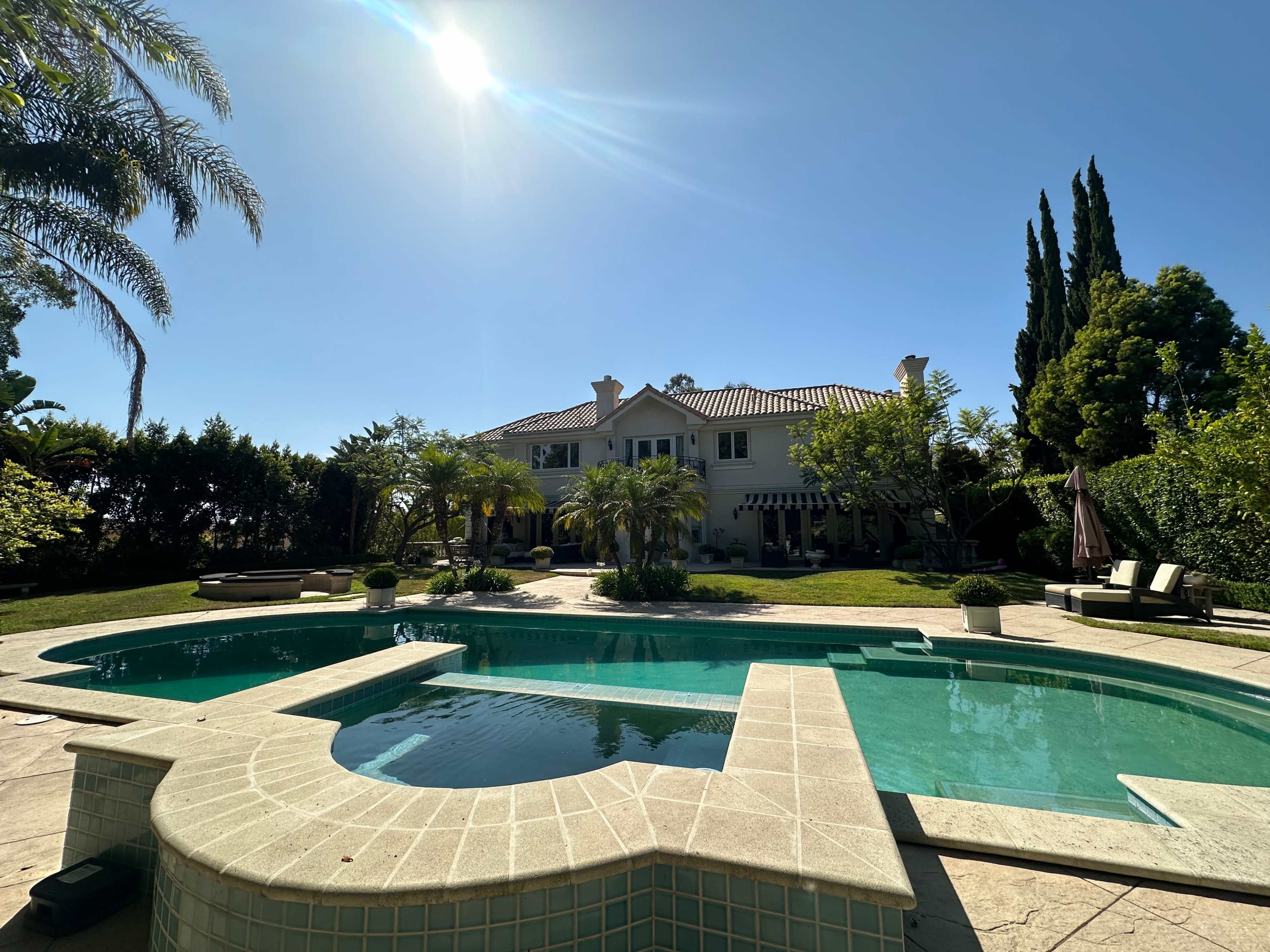A large, modern house with a swimming pool and landscaped garden is surrounded by palm trees under a clear blue sky.