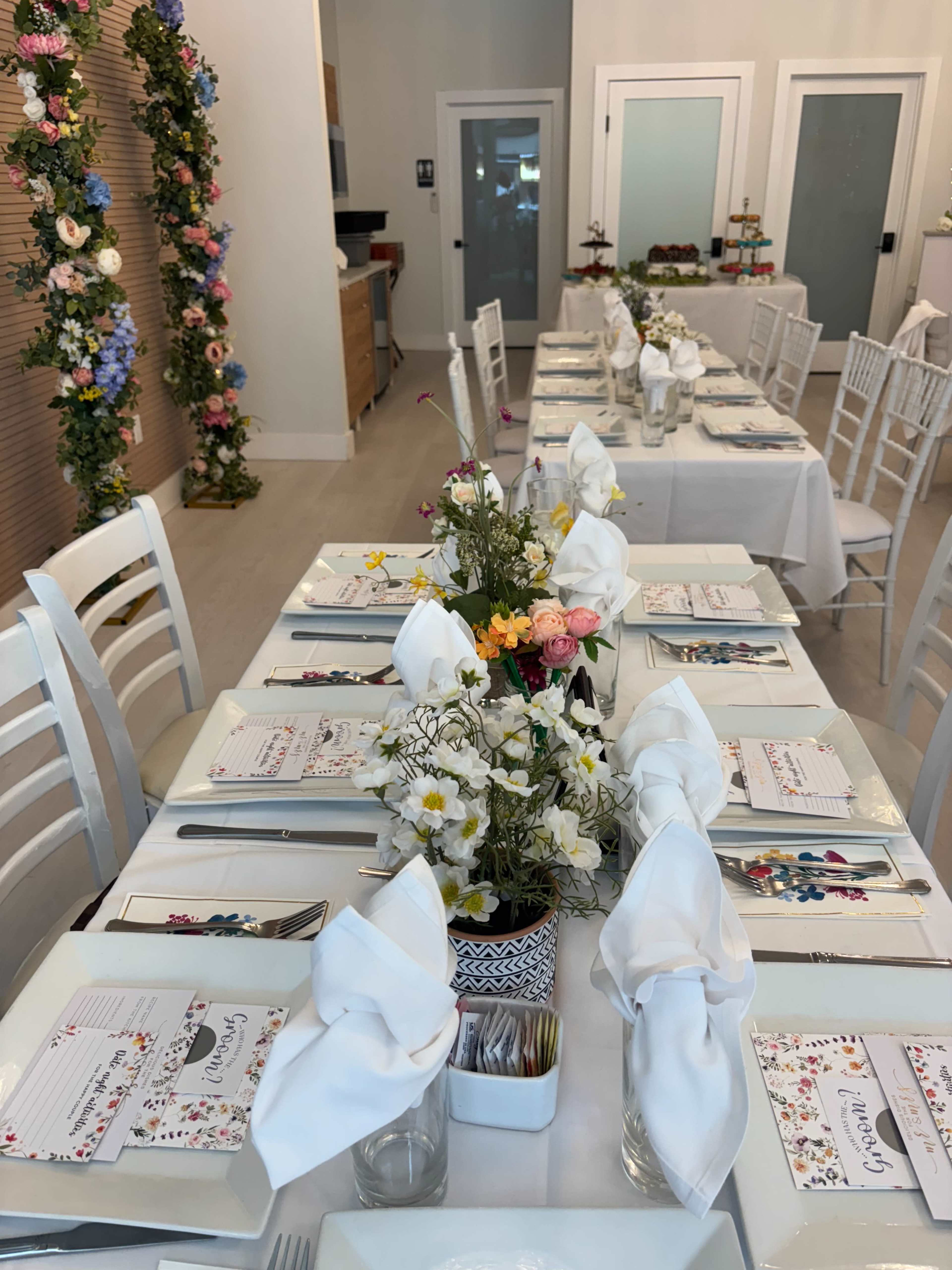 A rectangular dining table is set with white tablecloths, elegant dinnerware, and floral centerpieces in a well-lit room decorated with colorful flowers.
