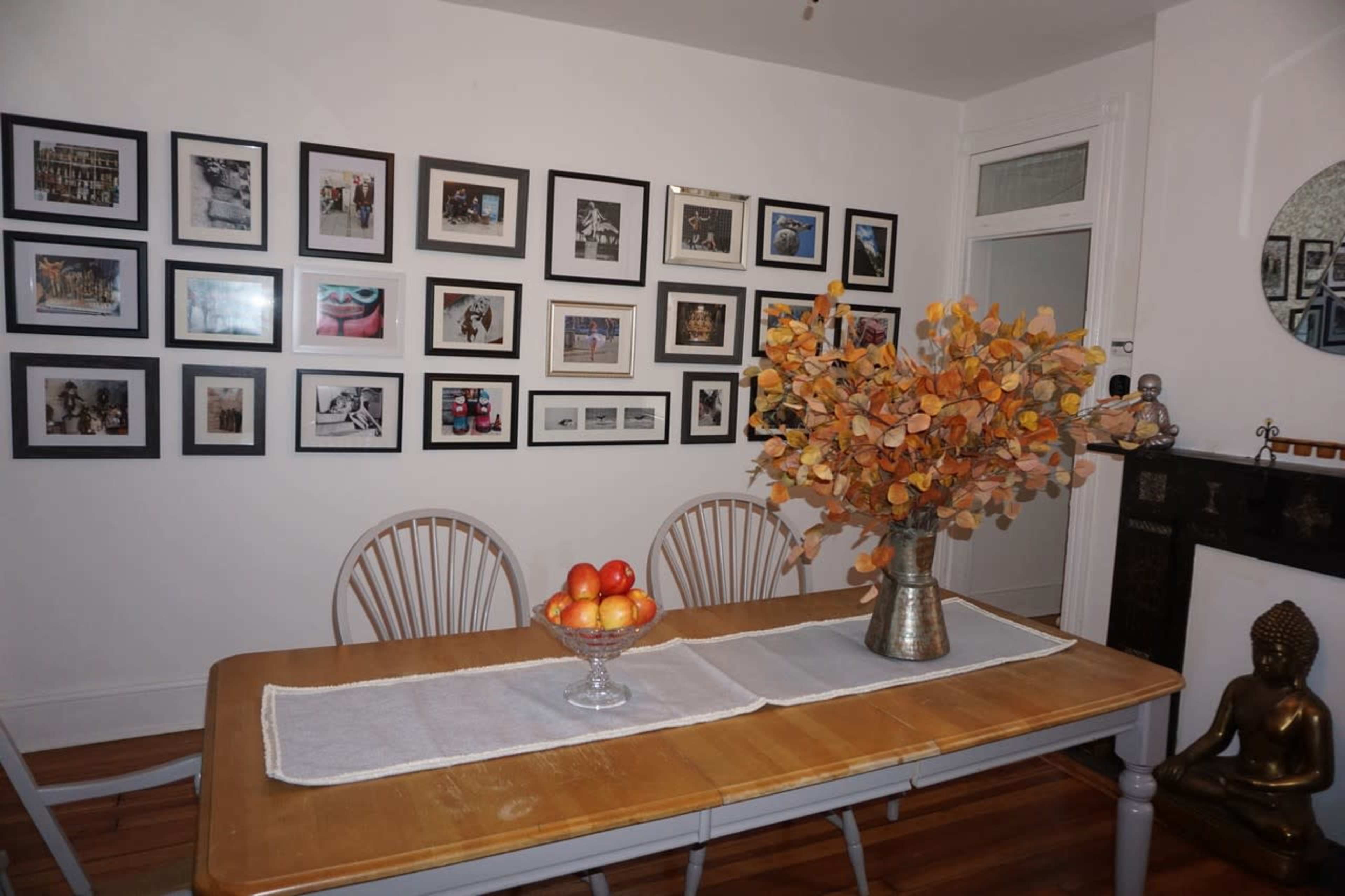 A dining room features a wooden table with a vase of orange leaves and a bowl of apples, surrounded by framed photographs on the wall.