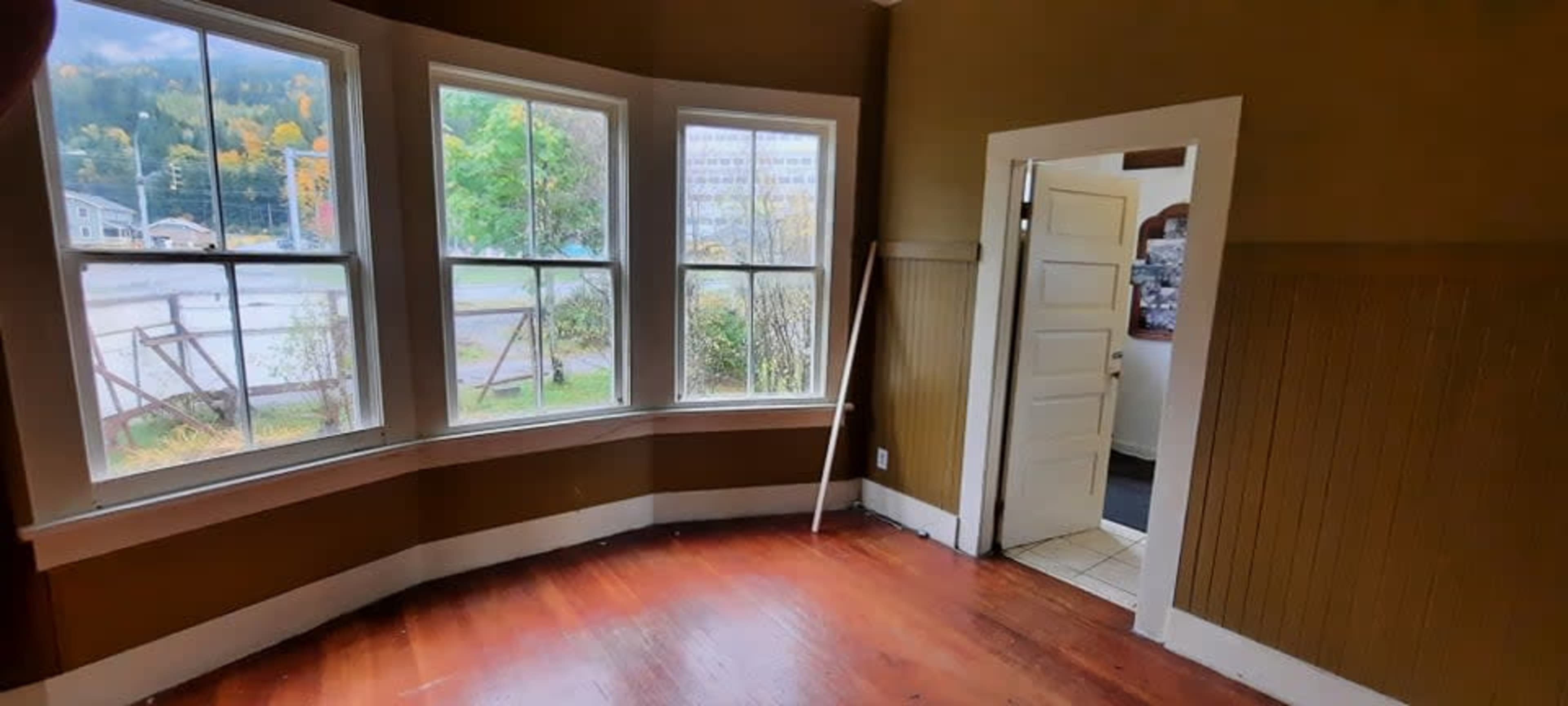 The image shows an empty room with three large windows and a door leading to another space, featuring wooden floors and brown walls.