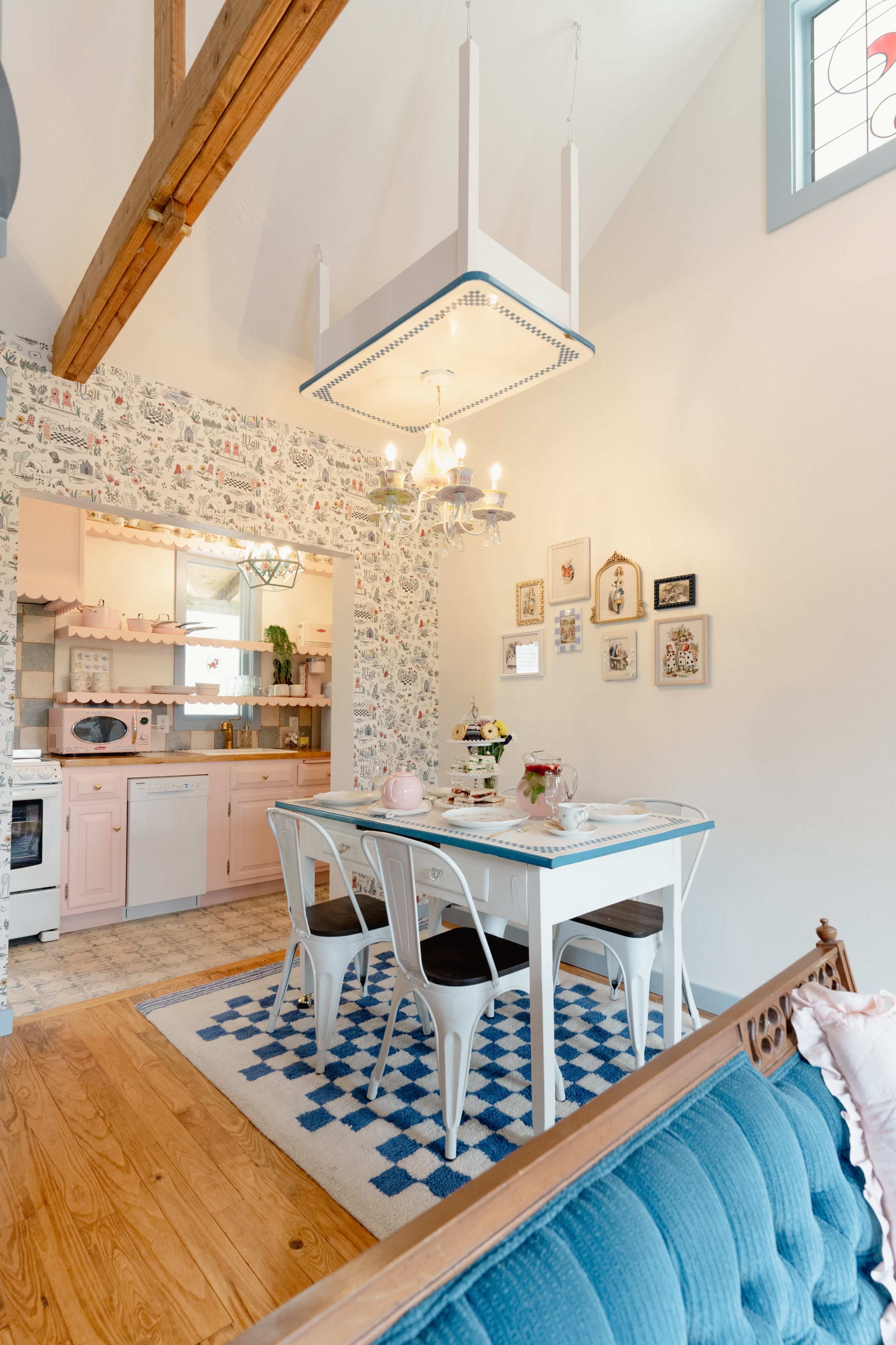 The scene depicts a bright dining area featuring a white table with blue chairs, set against a backdrop of floral wallpaper and a modern chandelier.