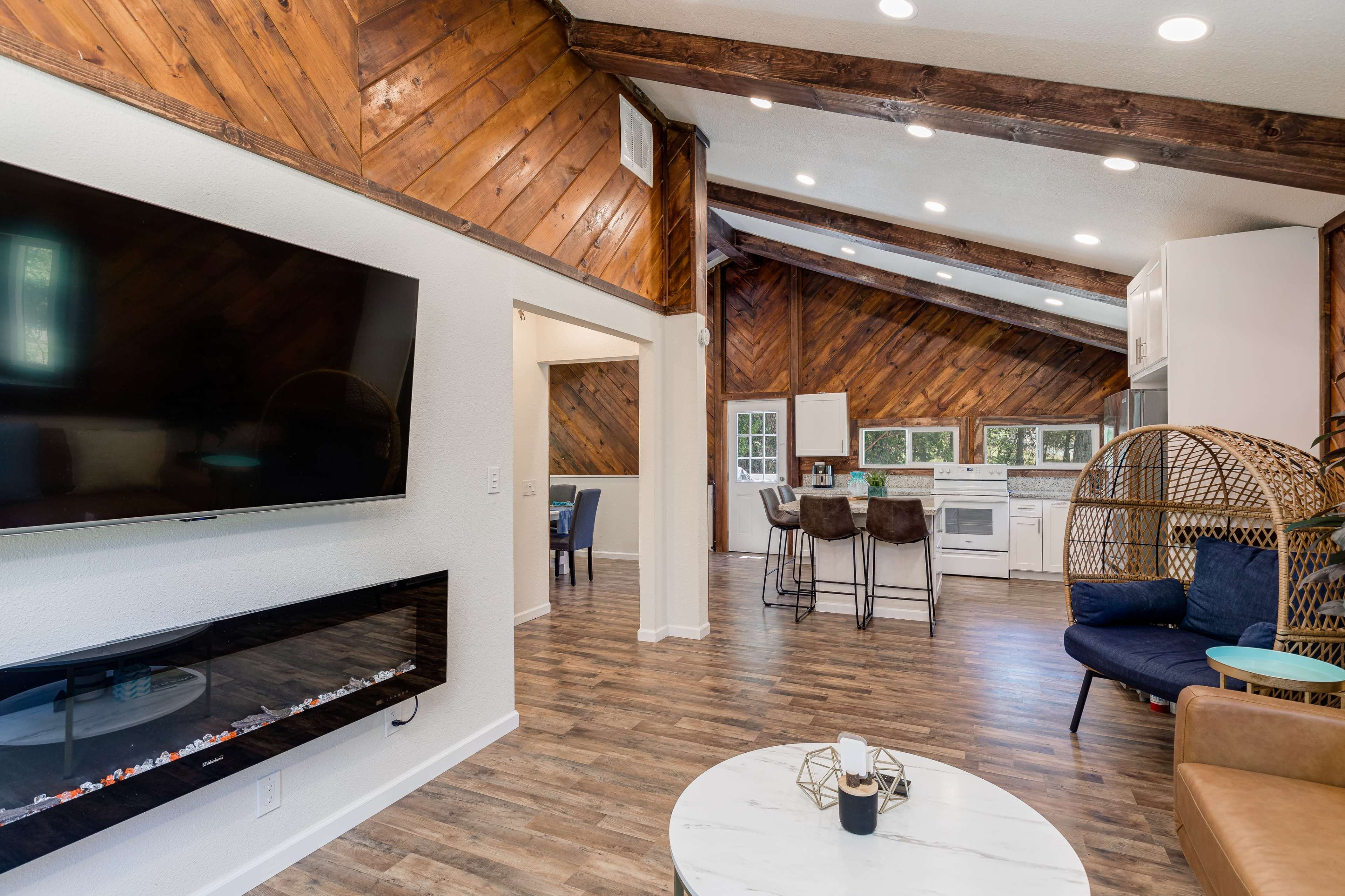A modern living room with a flat-screen TV mounted on the wall, a contemporary electric fireplace, and an open kitchen area featuring white cabinetry and a wooden ceiling.