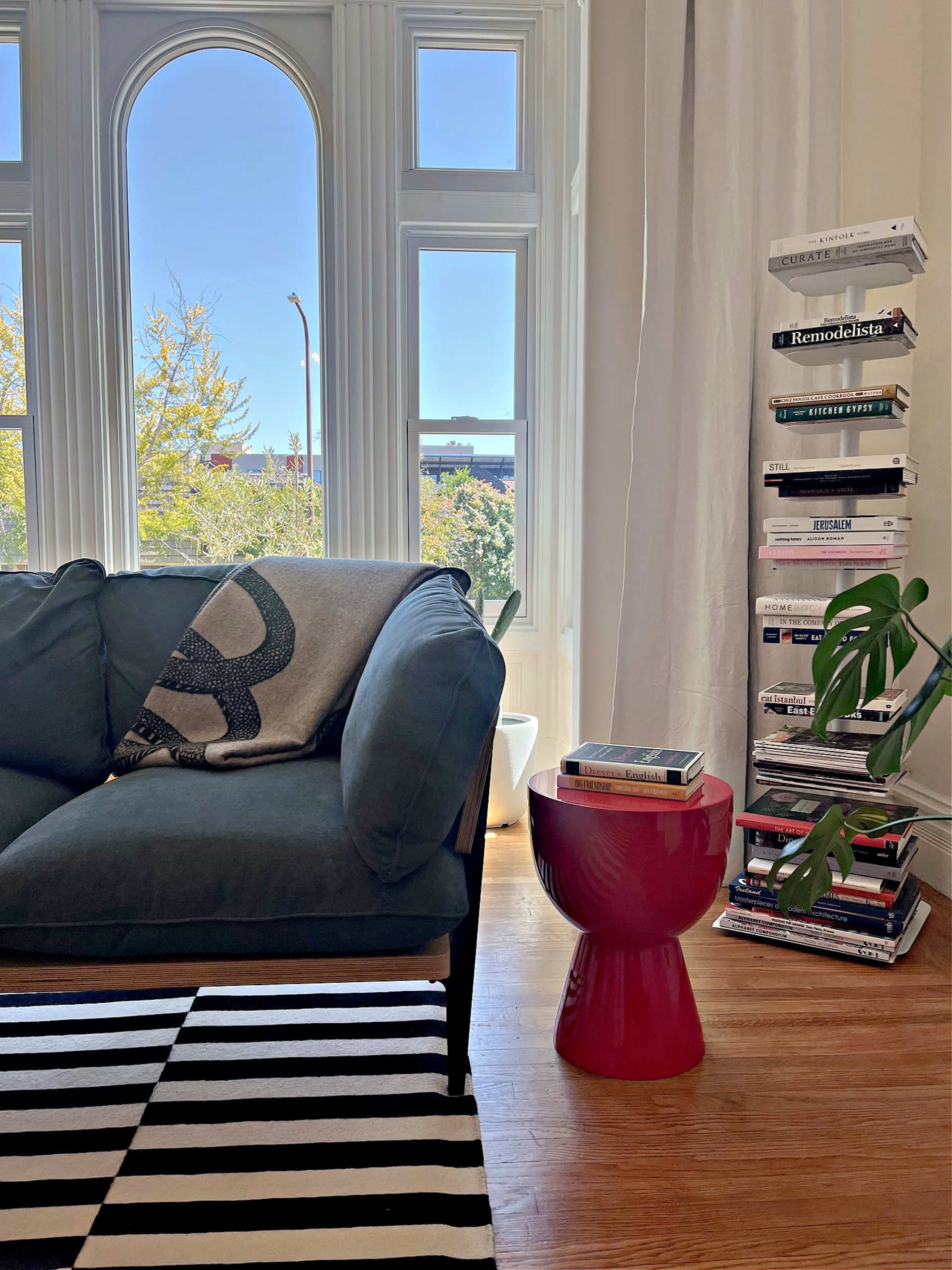 A living room features a dark green sofa, a striped rug, a red side table, and a stack of books beside a window with a view of trees.